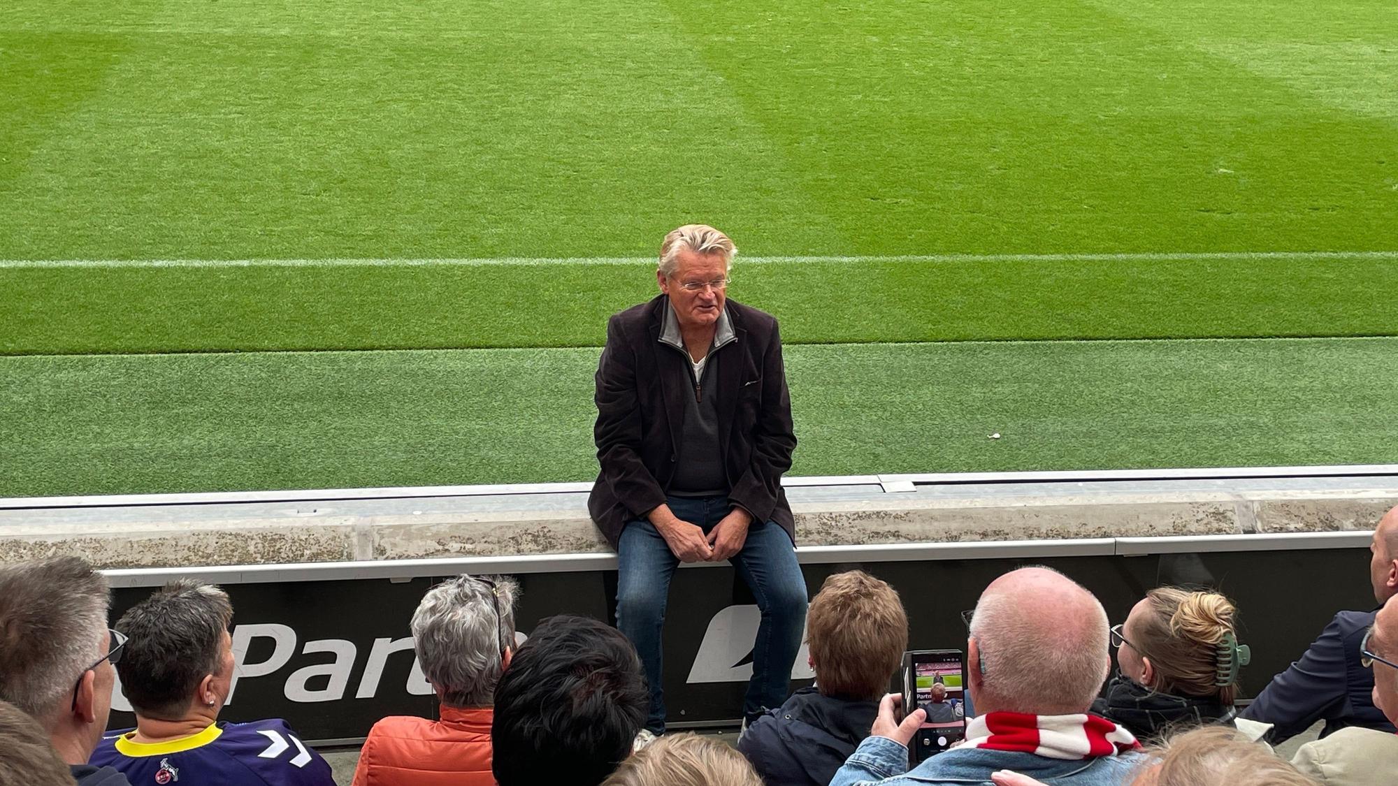 Ein Mann sitzt am Rand der Tribünenbegrenzung im Stadion und spricht mit den Menschen, die vor ihm sitzen.