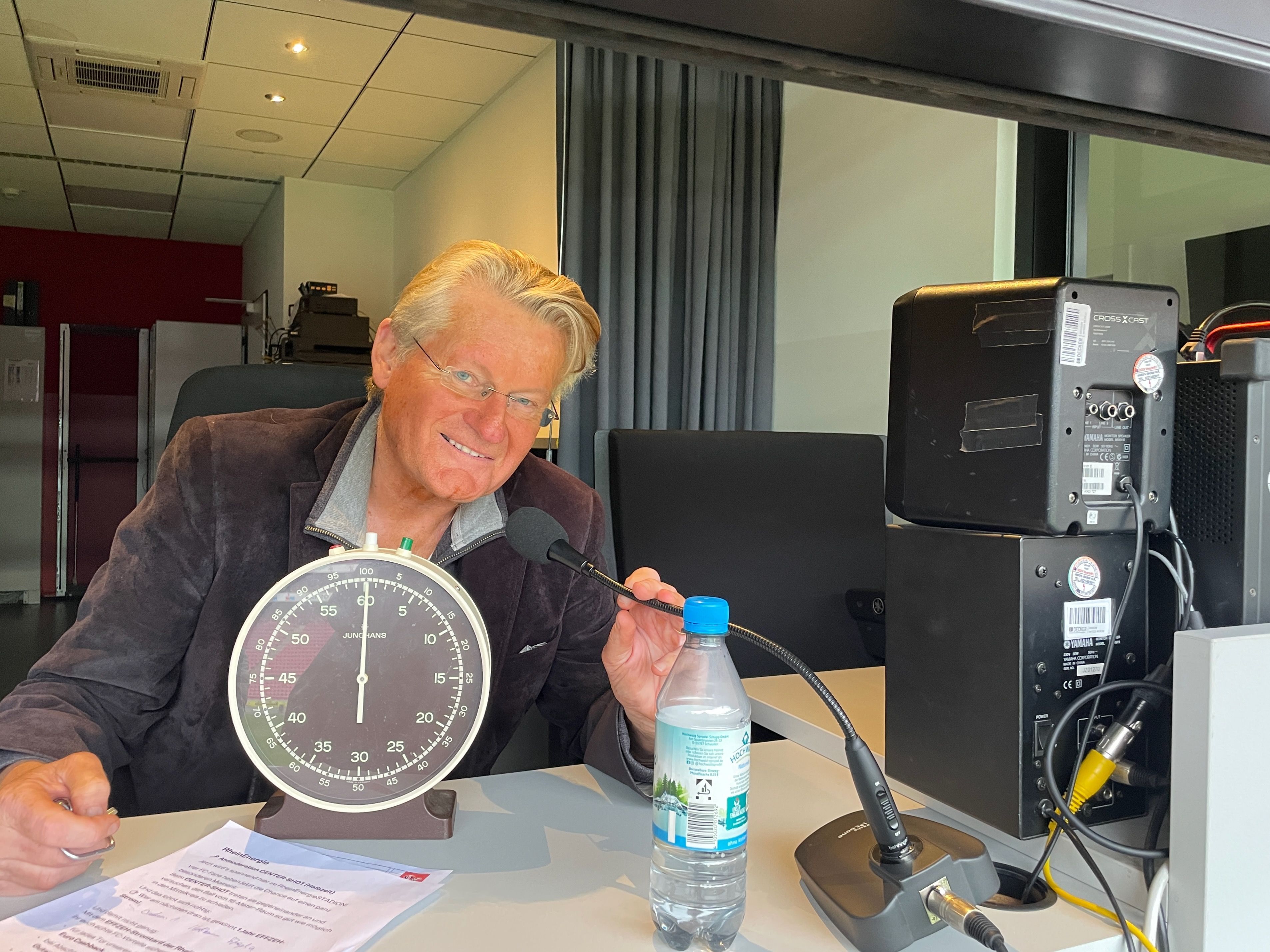 Ein Mann im Jackett sitzt hinter einer großen Stoppuhr an einem Tisch, auf dem auch ein Mikrofon steht.
