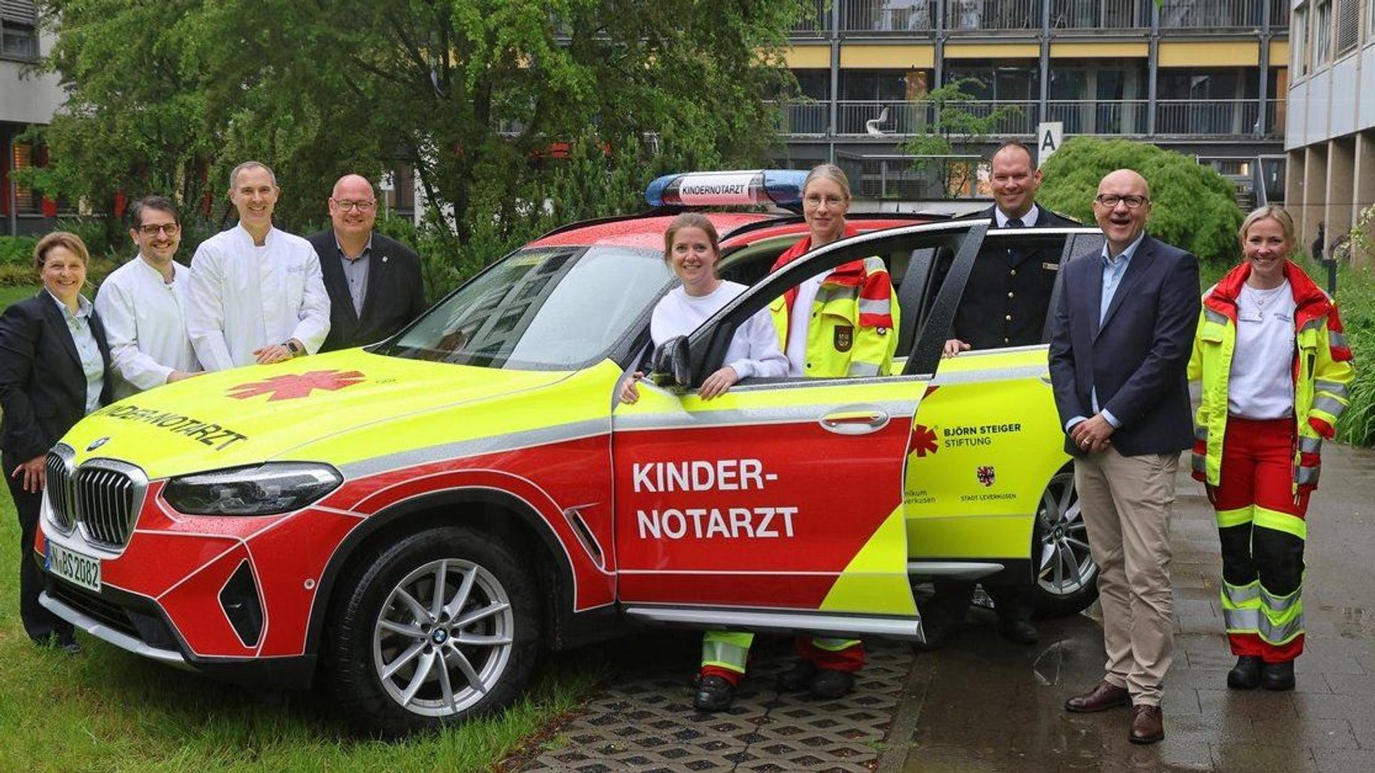 Ein Notarztwagen in Rot und Neongelb, auf der Tür steht Kindernotarzt, drei Personen in Rettungssanitäter-Kleidung und zwei Ärzte in Weiß, weitere drei Personen aus der Verwaltung und ein Mann von der Feuerwehr