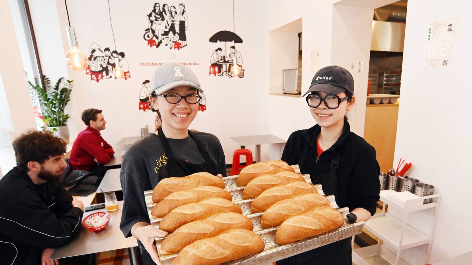 Die beiden Frauen zeigen auf einem großen Tablett die Baguettbrötchen. Beide tragen Baseball Caps und lachen in die Kamera.