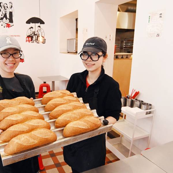 Die beiden Frauen zeigen auf einem großen Tablett die Baguettbrötchen. Beide tragen Baseball Caps und lachen in die Kamera.