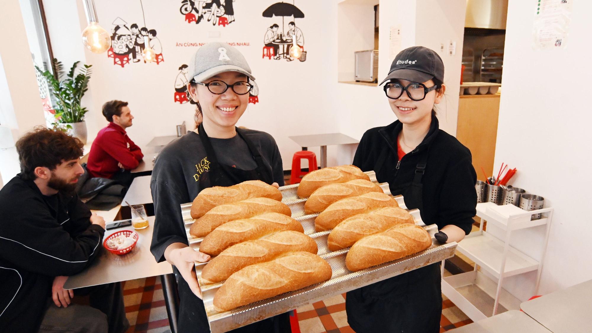 Die beiden Frauen zeigen auf einem großen Tablett die Baguettbrötchen. Beide tragen Baseball Caps und lachen in die Kamera.
