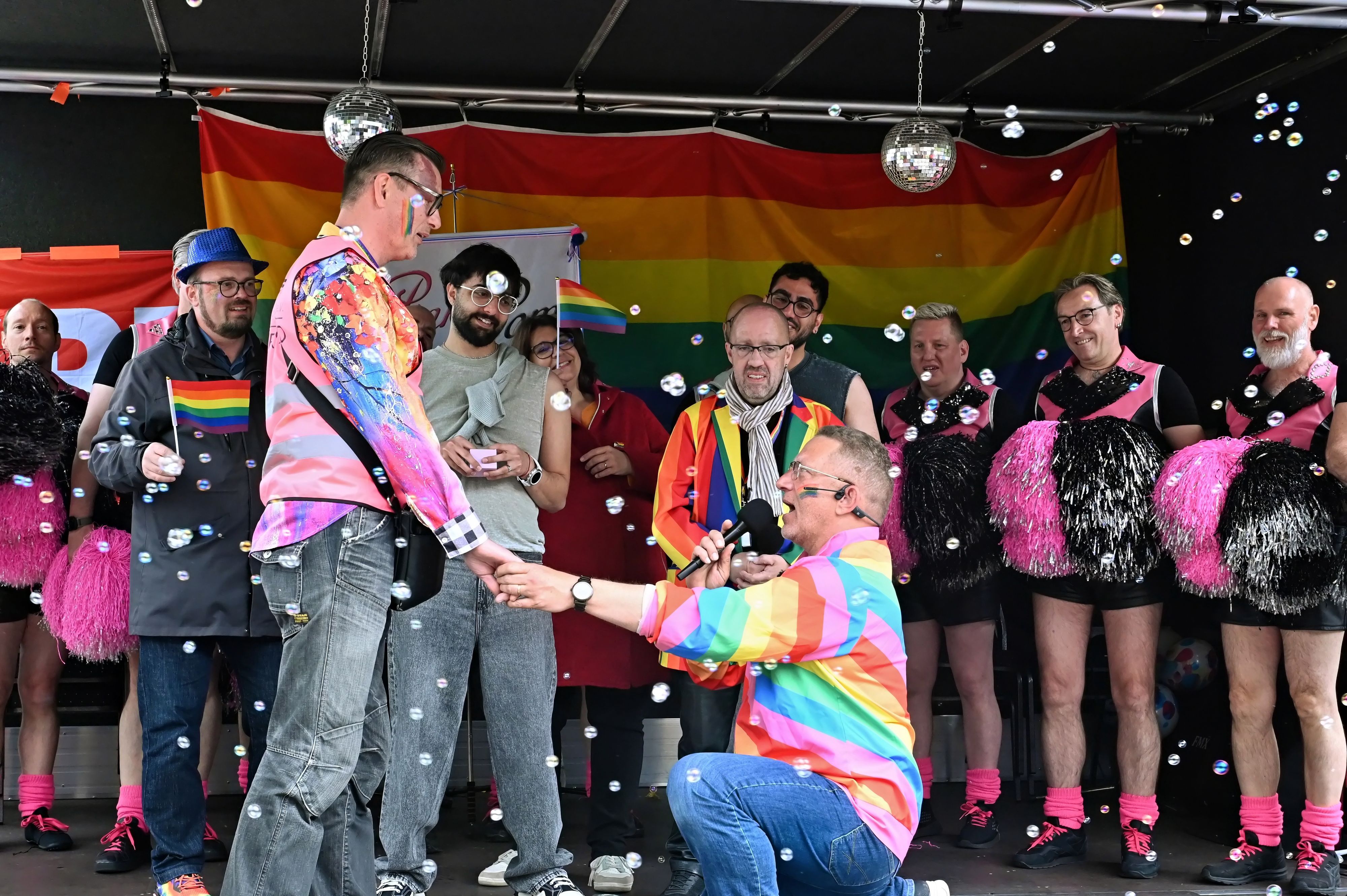 Winfried Kubitza-Simons kniet vor seinem Mann Mario Kubitza, hält dessen Hand und macht ihm einen Antrag. Im Hintergrund sind männliche Cheerleader in schwarz-rosa-farbenen Kostümen zu sehen.