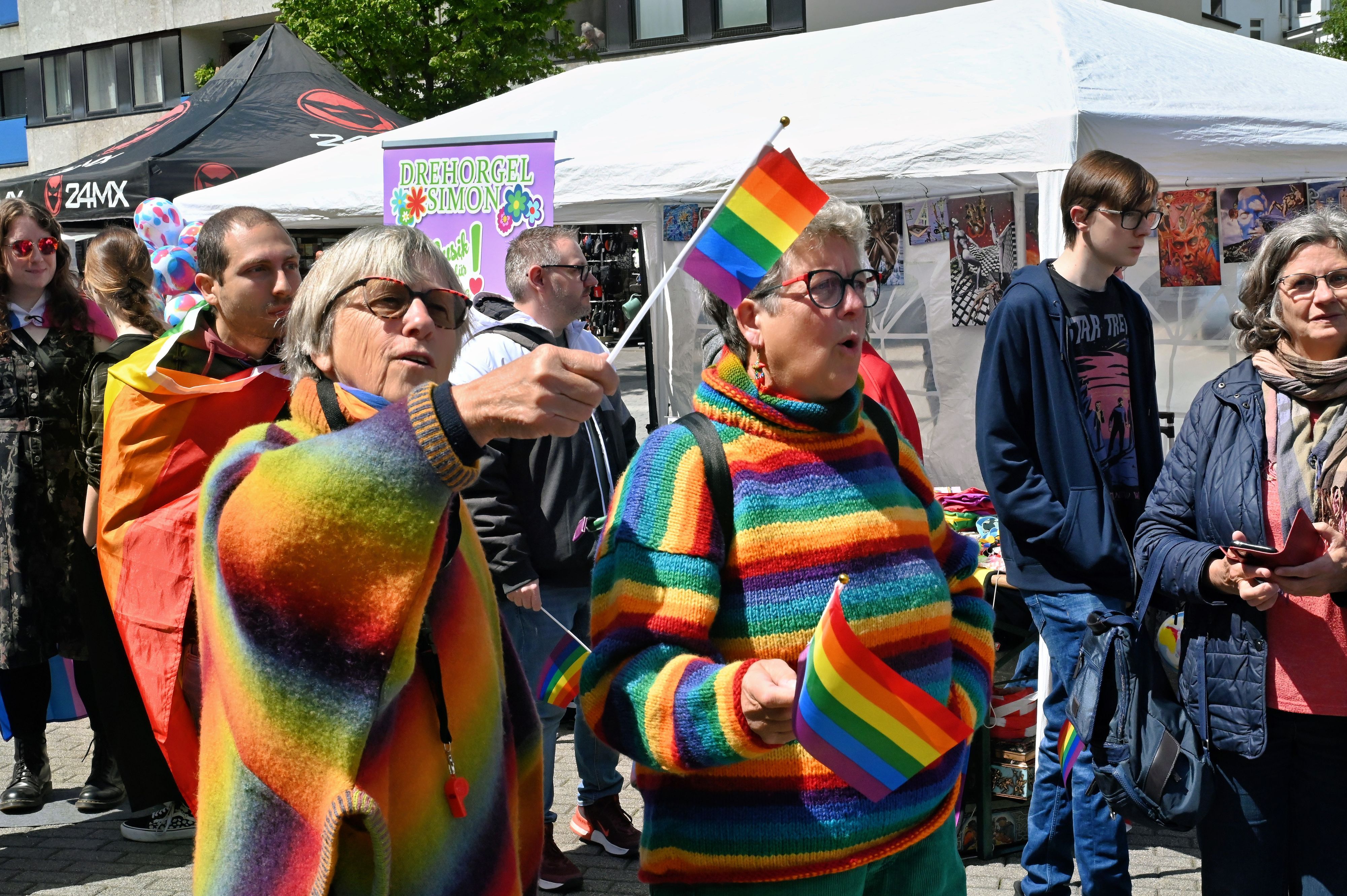 Zwei farbenfroh gekleidete Frauen halten kleine Fähnchen mit den Regenbogenfarben.
