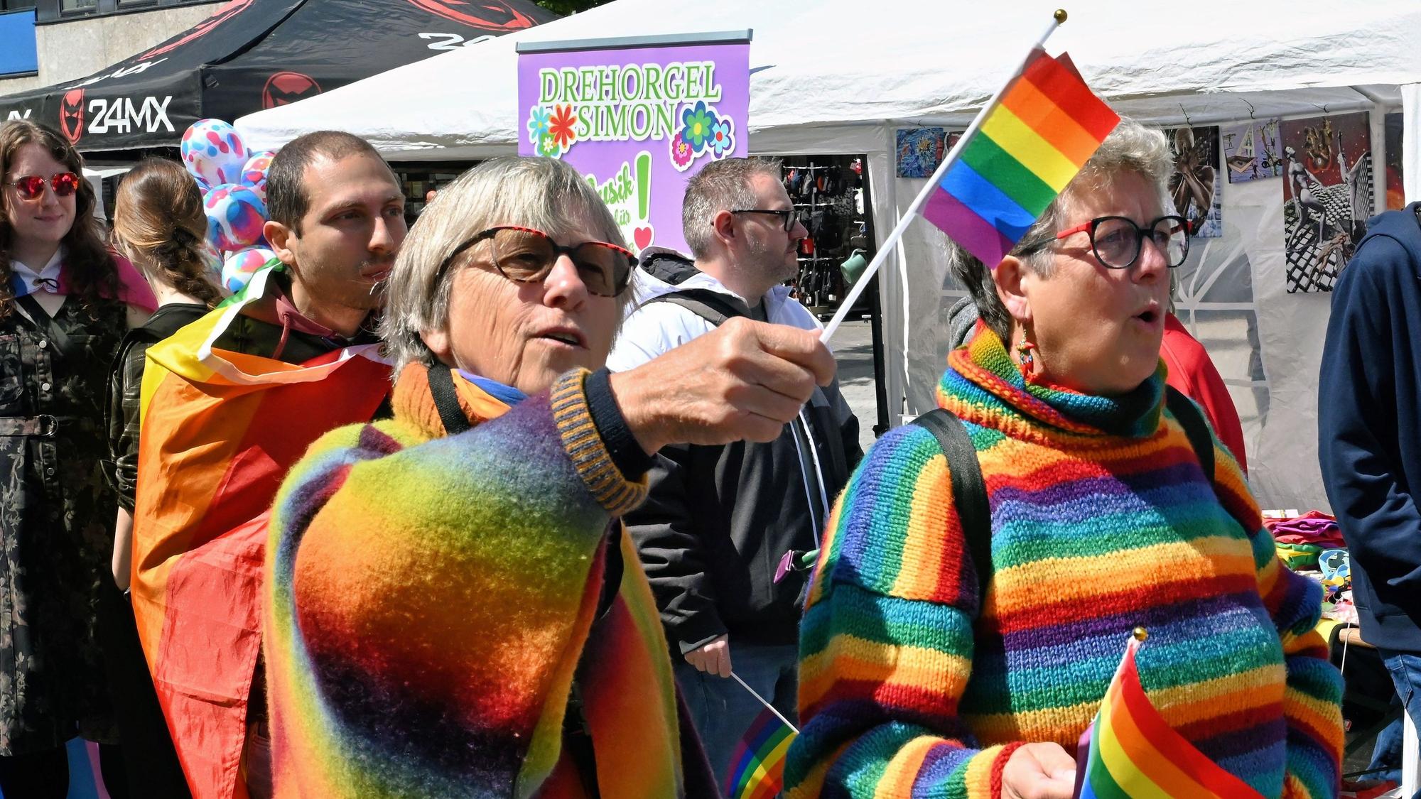 Zwei farbenfroh gekleidete Frauen halten kleine Fähnchen mit den Regenbogenfarben.