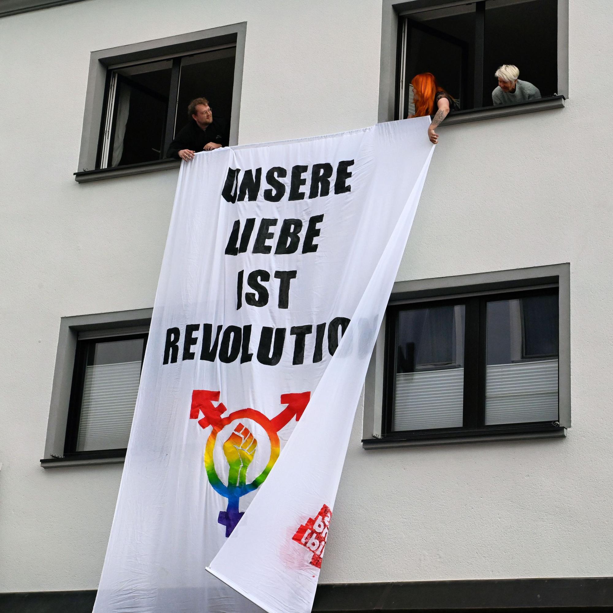Hausbewohner hängen aus Fenstern im Obergeschoss eine Fahne mit der Aufschrift „Unsere Liebe ist Revolution“.