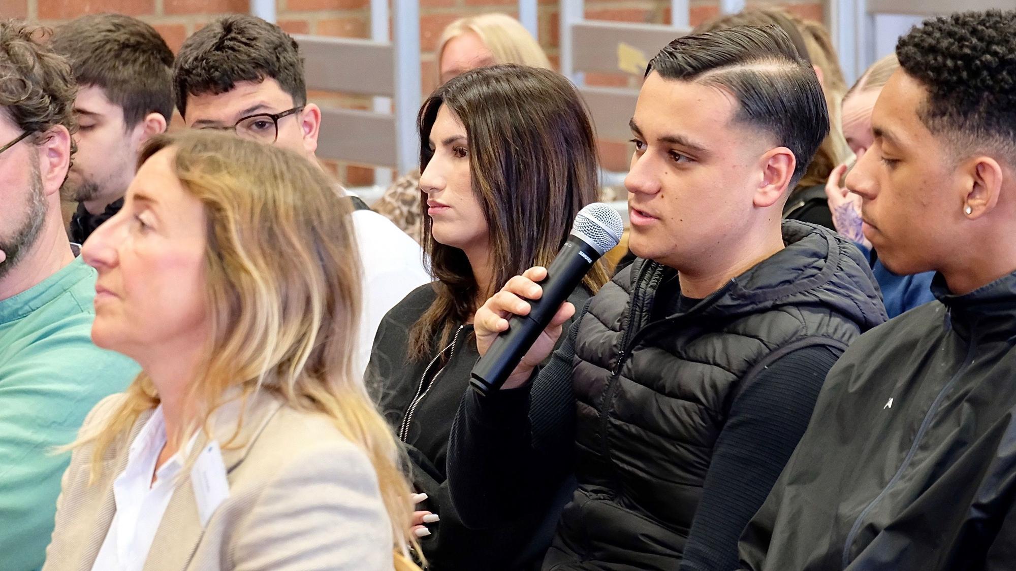 Schülerinnen und Schüler sitzen in Stuhlreihen. Einer von ihnen spricht in ein Mikrofon.