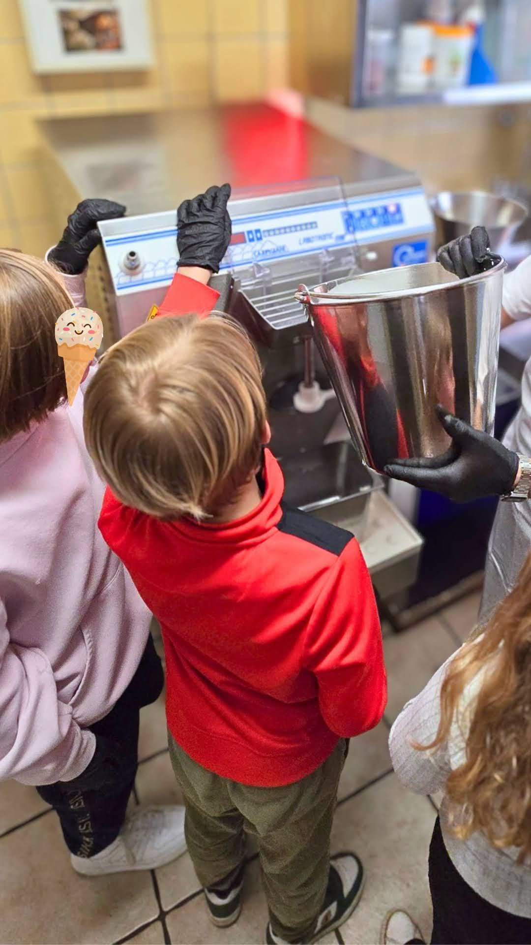 Zwei Kinder stehen vor einer Eismaschine. Ein Erwachsener hält einen Metalleimer, in den das Eis kommt.