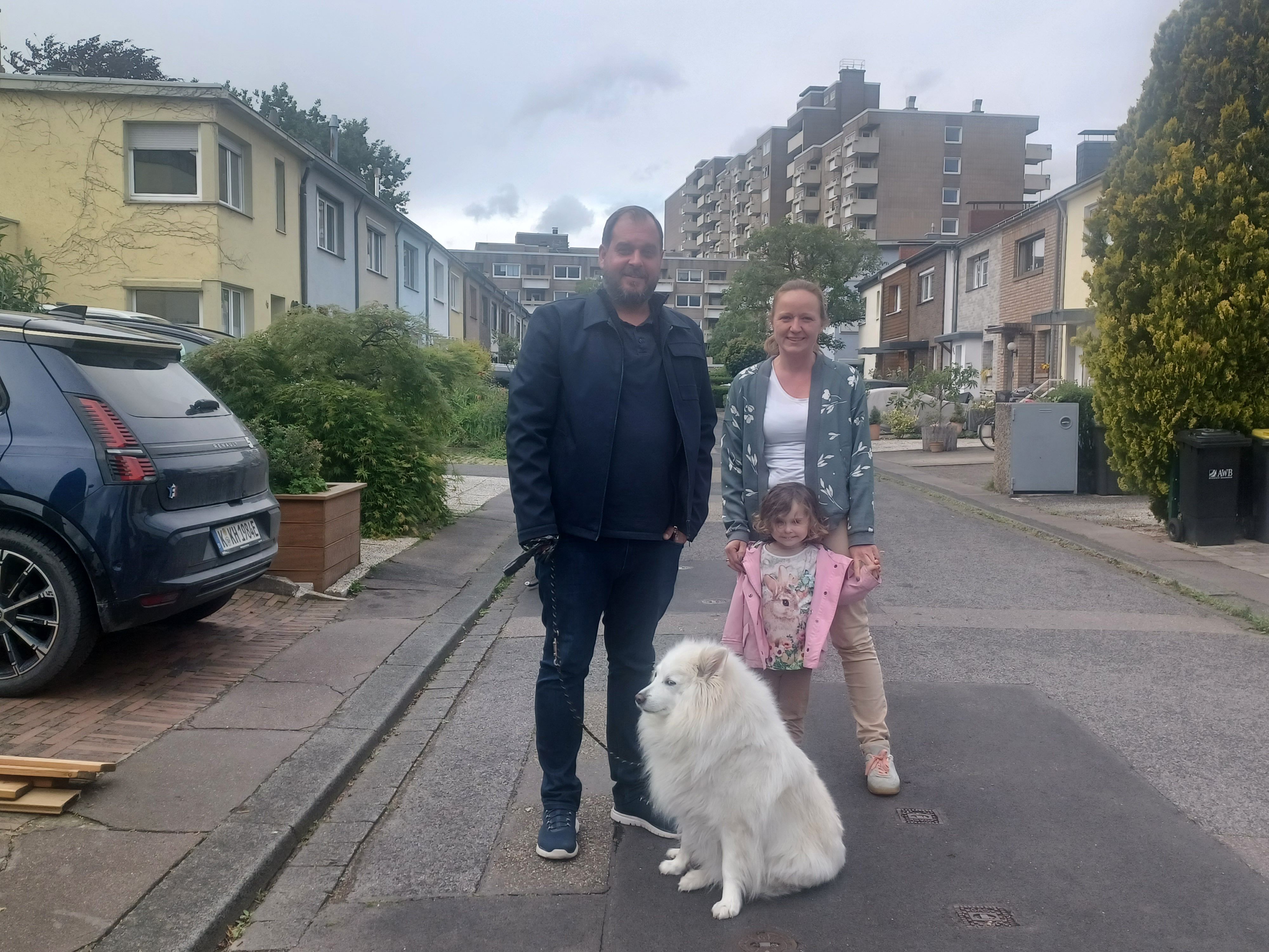 Ein Mann, eine Frau, ein Kind und ein Hund stehen auf einer schmalen Anwohnerstraße.
