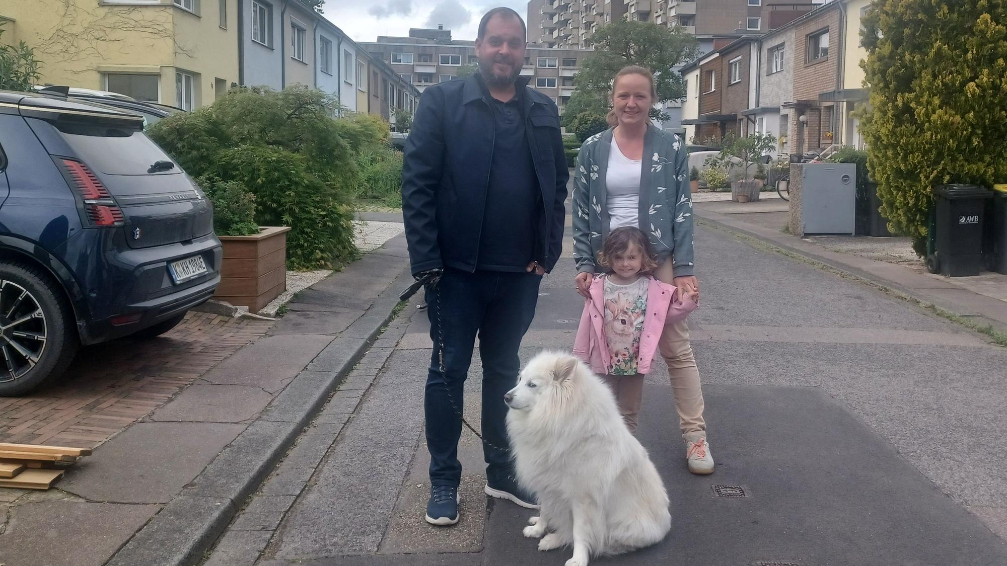 Ein Mann, eine Frau, ein Kind und ein Hund stehen auf einer schmalen Anwohnerstraße.