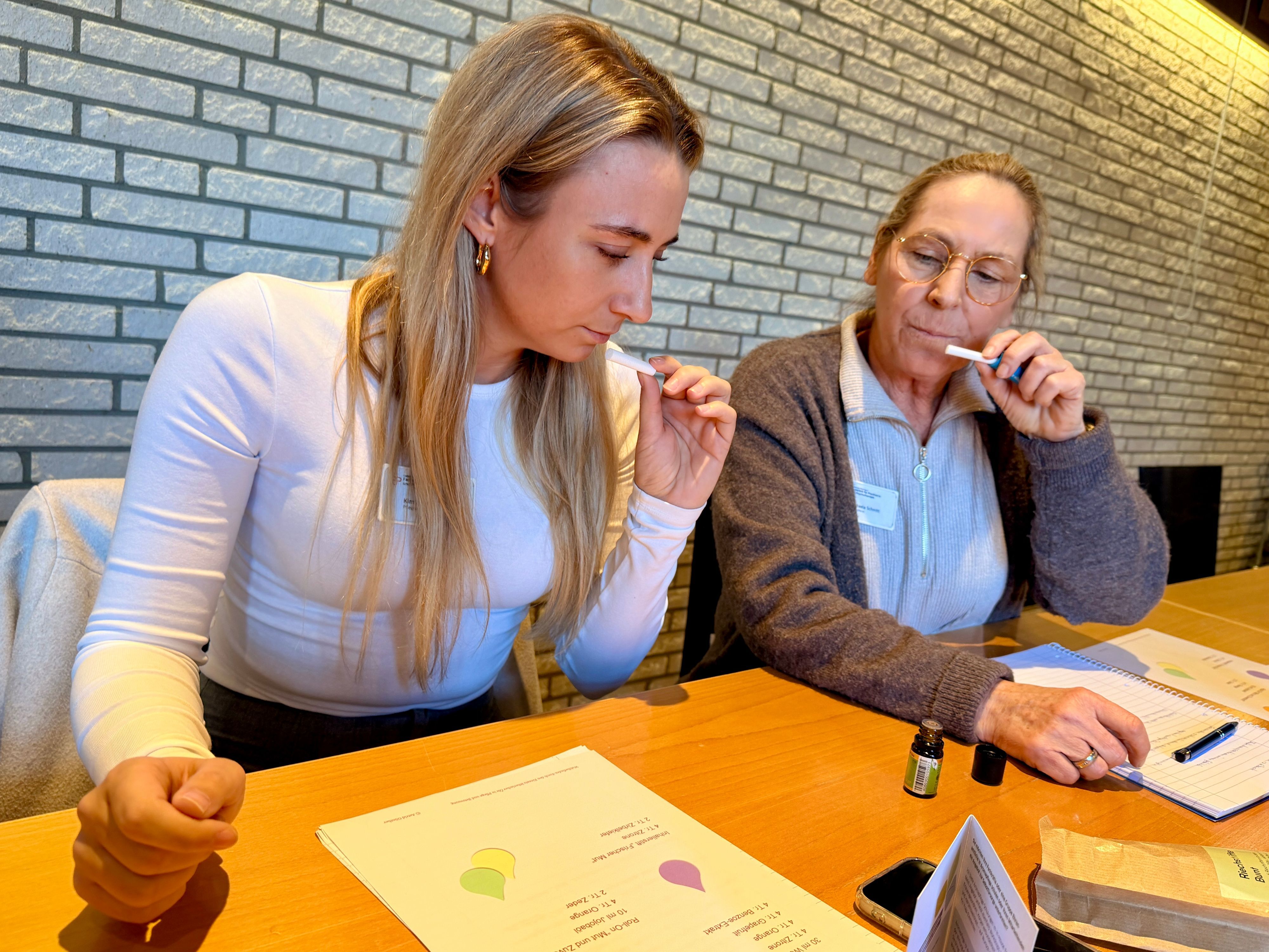Zwei Frauen halten sich schnuppernd kleine Papierstreifen vor die Nase. Auf dem Tisch steht ein aufgeschraubtes Glasfläschchen.