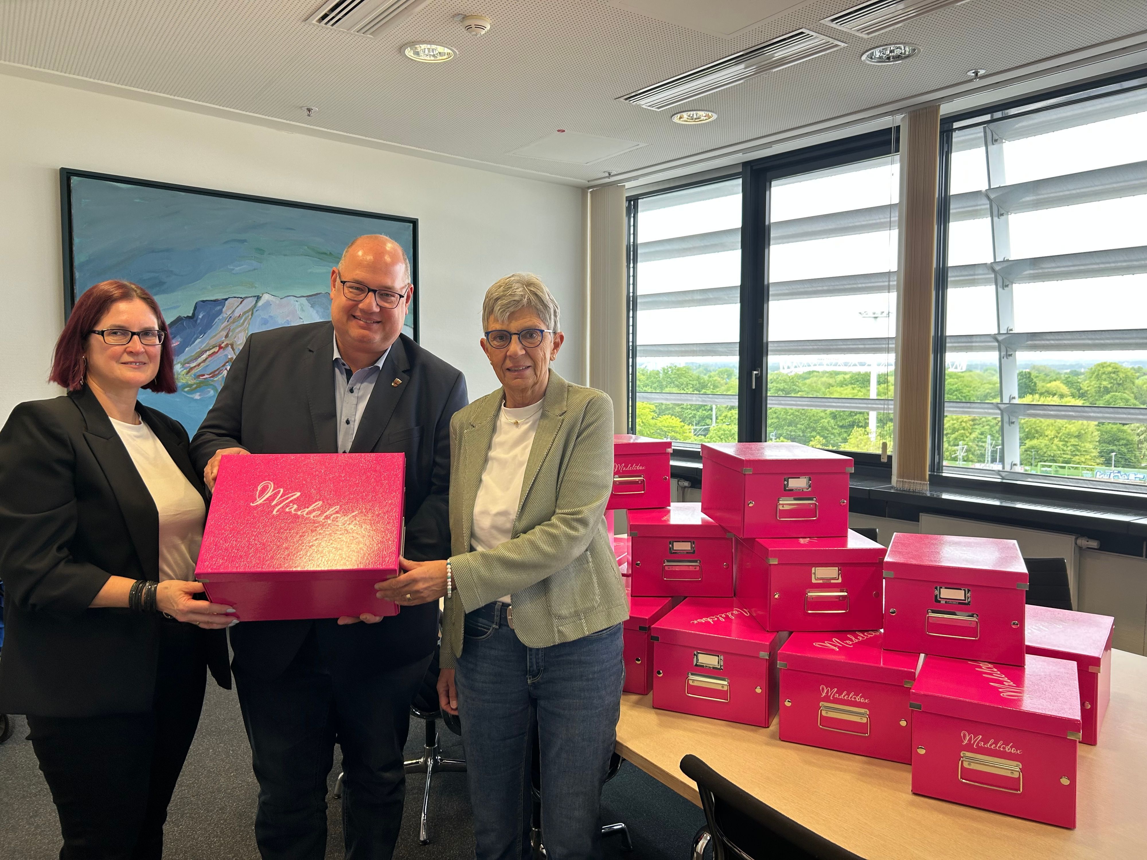 Zwei Frauen und ein Mann halten einen rosafarbenen Karton hoch.