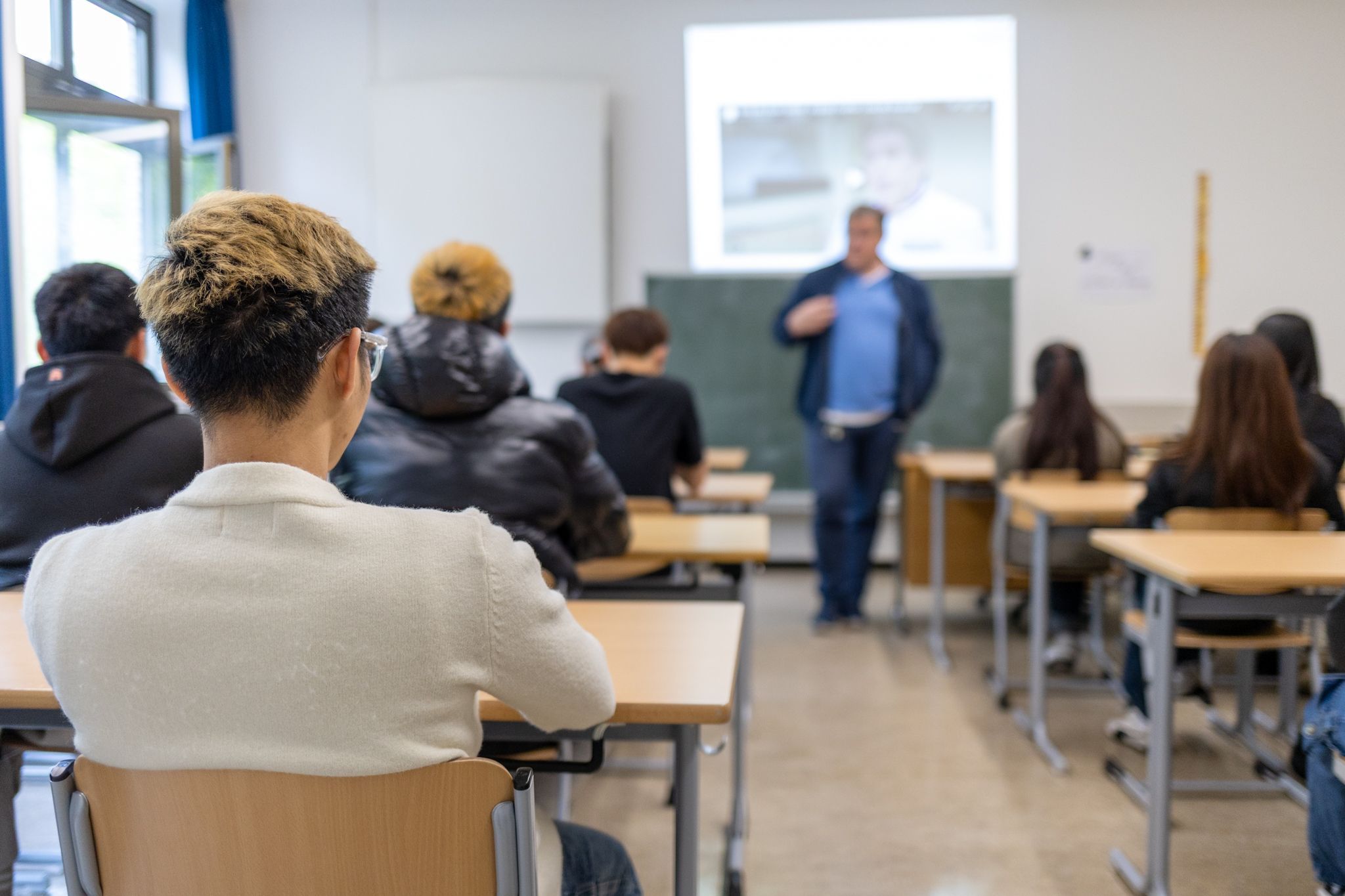 Eine Schulklasse im Hintergrund, im Vordergrund ein Schüler von hinten abgebildet.