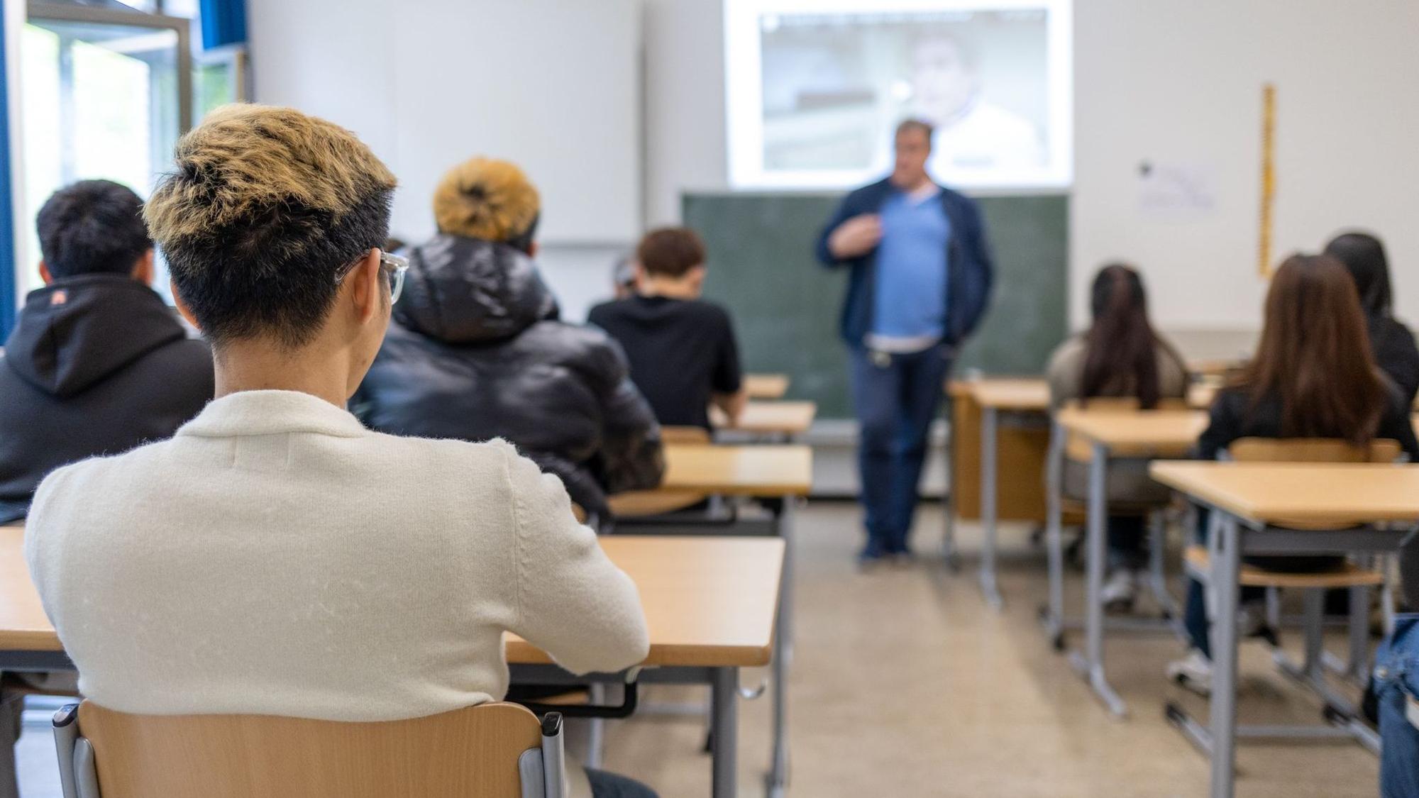 Eine Schulklasse im Hintergrund, im Vordergrund ein Schüler von hinten abgebildet.
