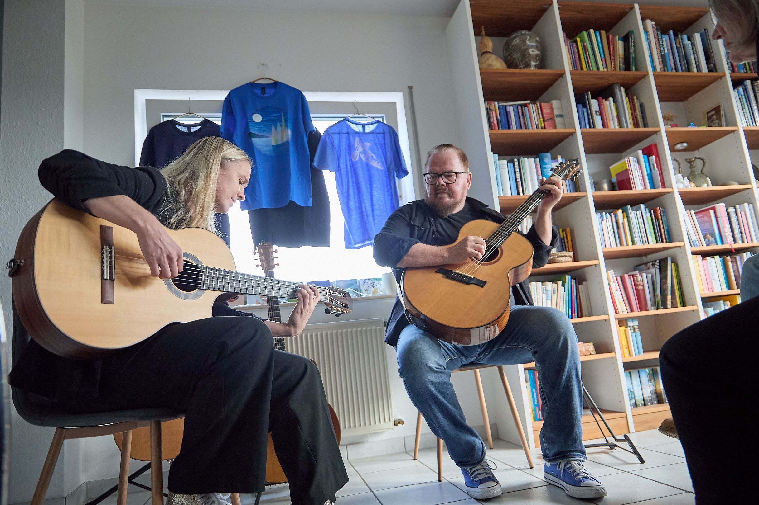 Ein Mann und eine Frau sitzen vor einem Bücherregal auf Stühlen und spielen auf Gitarren.