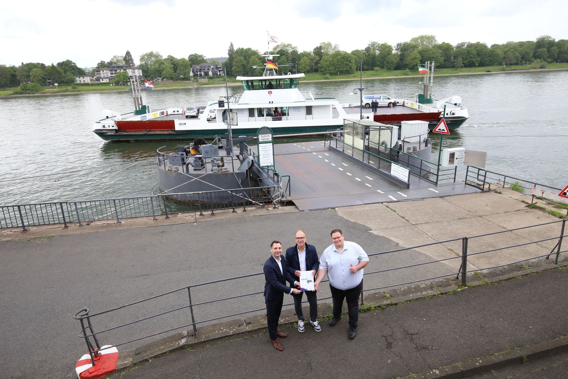 Drei Männer stehen vor der Rheinfähre Königswinter, die gerade abgelegt hat.