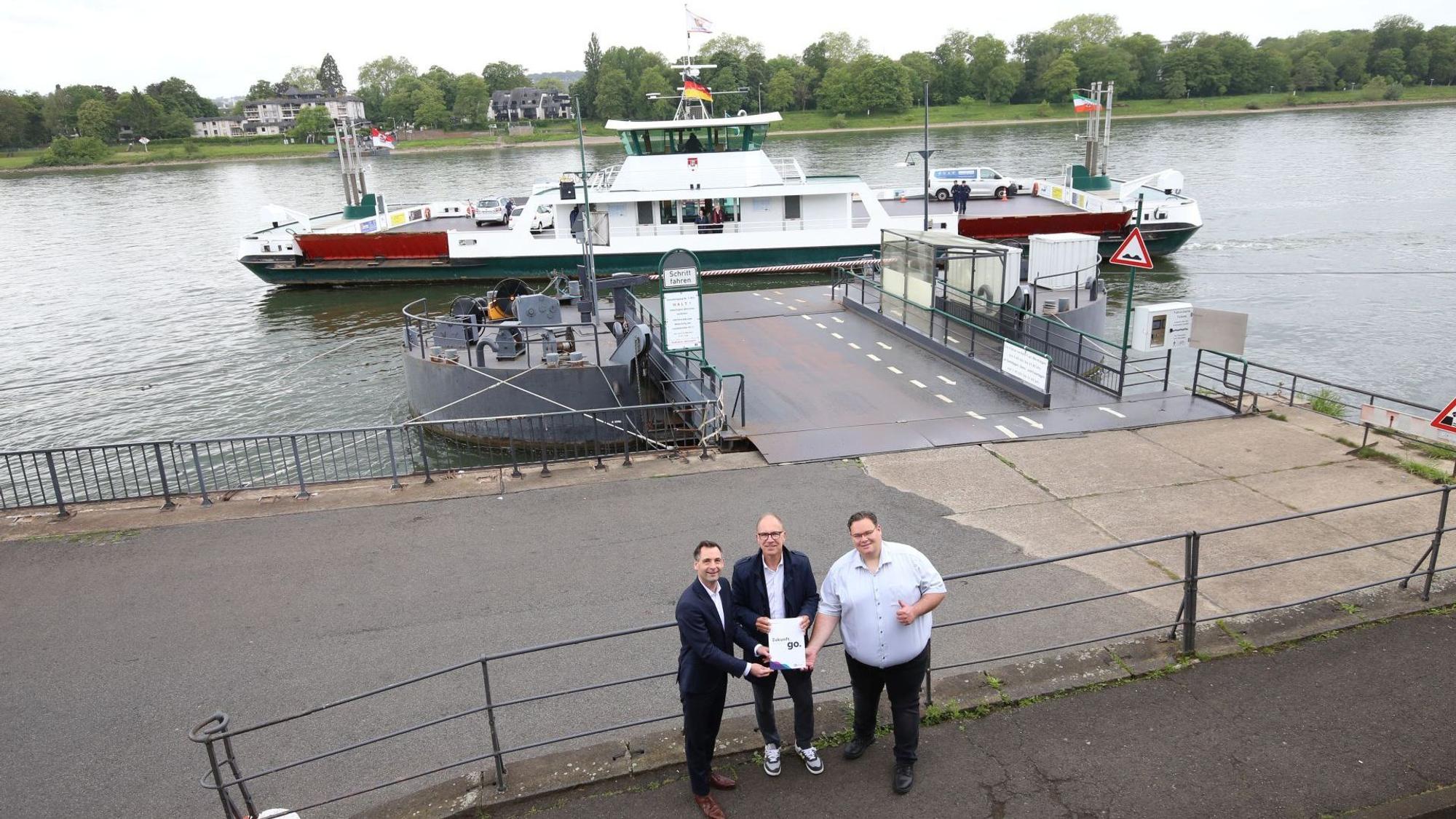 Drei Männer stehen vor der Rheinfähre Königswinter, die gerade abgelegt hat.