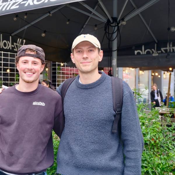 Zwei junge Männer stehen vor grünen Pflanzkübeln, sie haben Kappen auf, Pullis an.