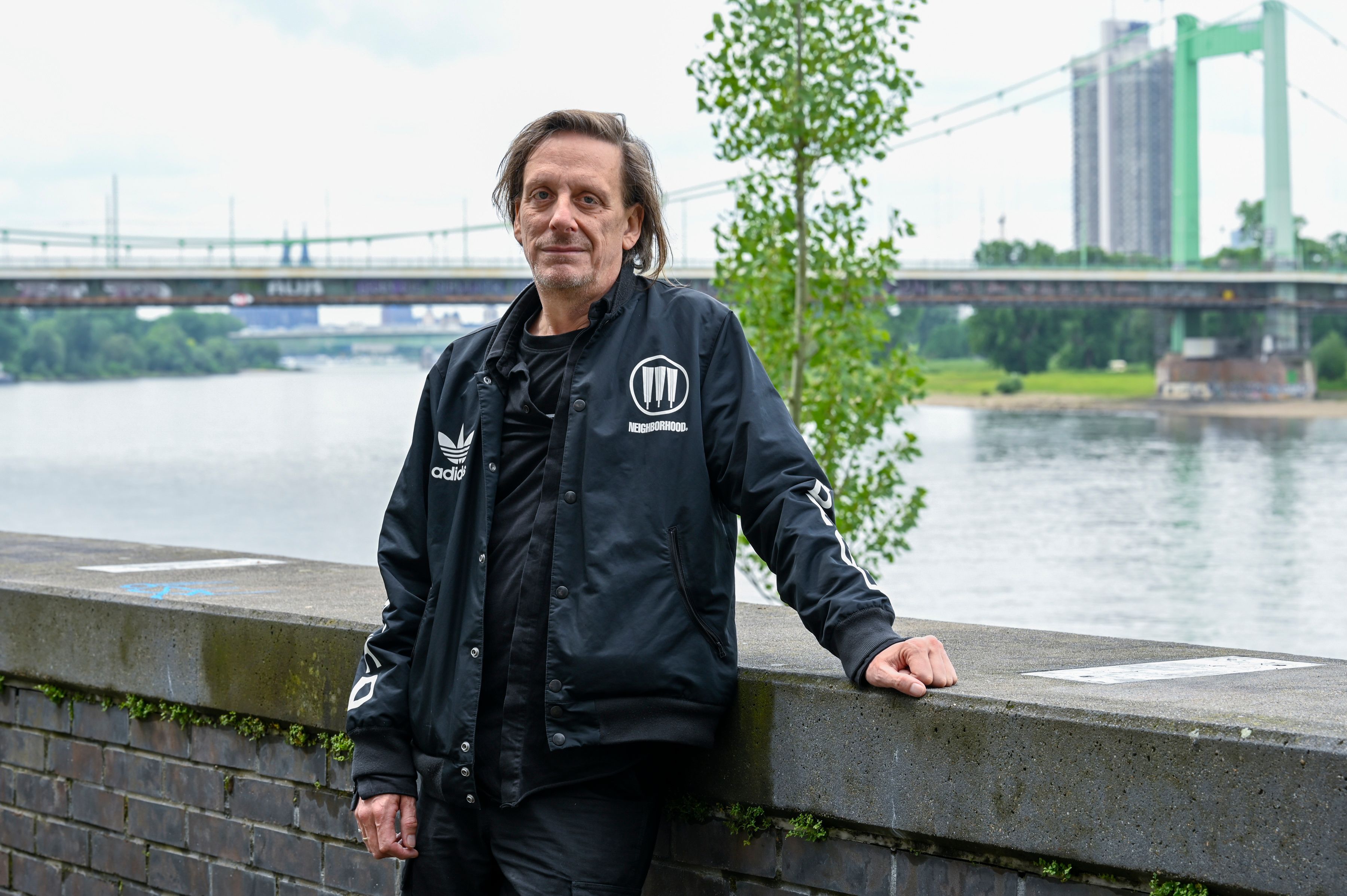 Ein Mann in schwarzen Klamotten steht vor einer Mauer am Rheinufer in Mülheim. Hinter ihm sind die Mülheimer Brücke und der Dom zu sehen.