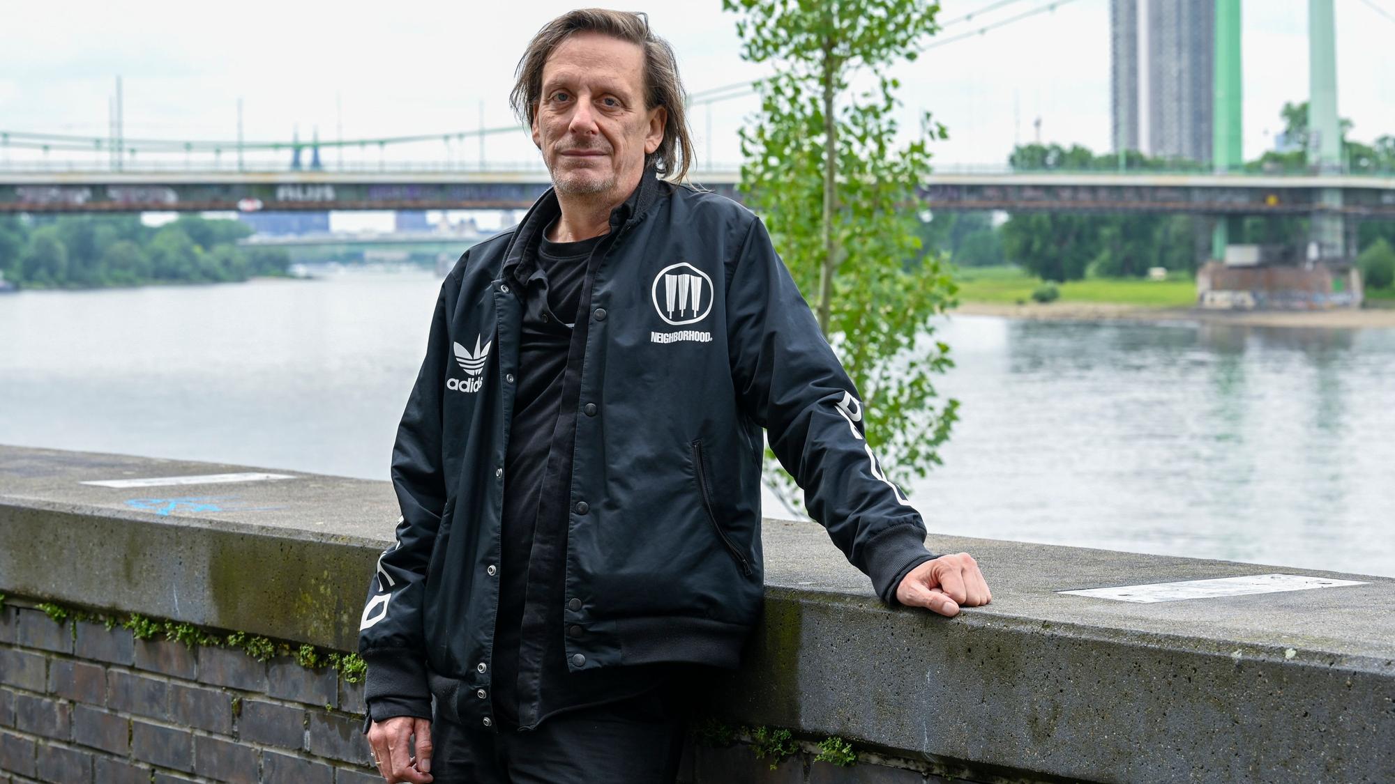 Ein Mann in schwarzen Klamotten steht vor einer Mauer am Rheinufer in Mülheim. Hinter ihm sind die Mülheimer Brücke und der Dom zu sehen.
