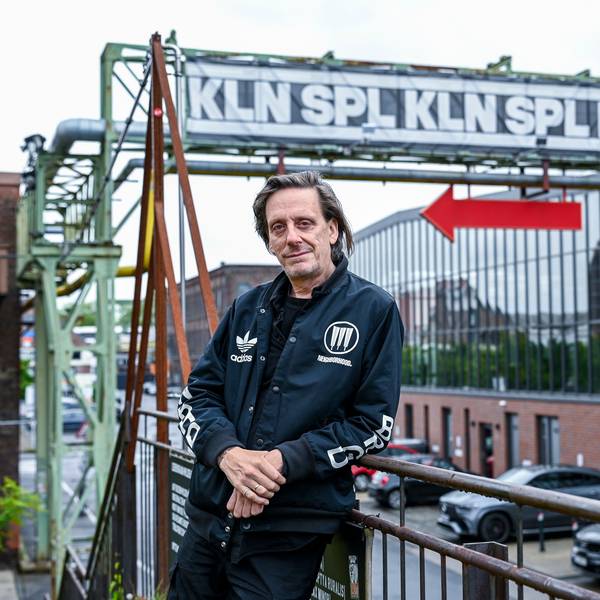 Ein Mann in schwarzen Klamotten steht vor einer Industrie-Kulisse. Über ihm der Schriftzug KLN SPL und ein roter Pfeil, der Richtung Eingang Depot weist.