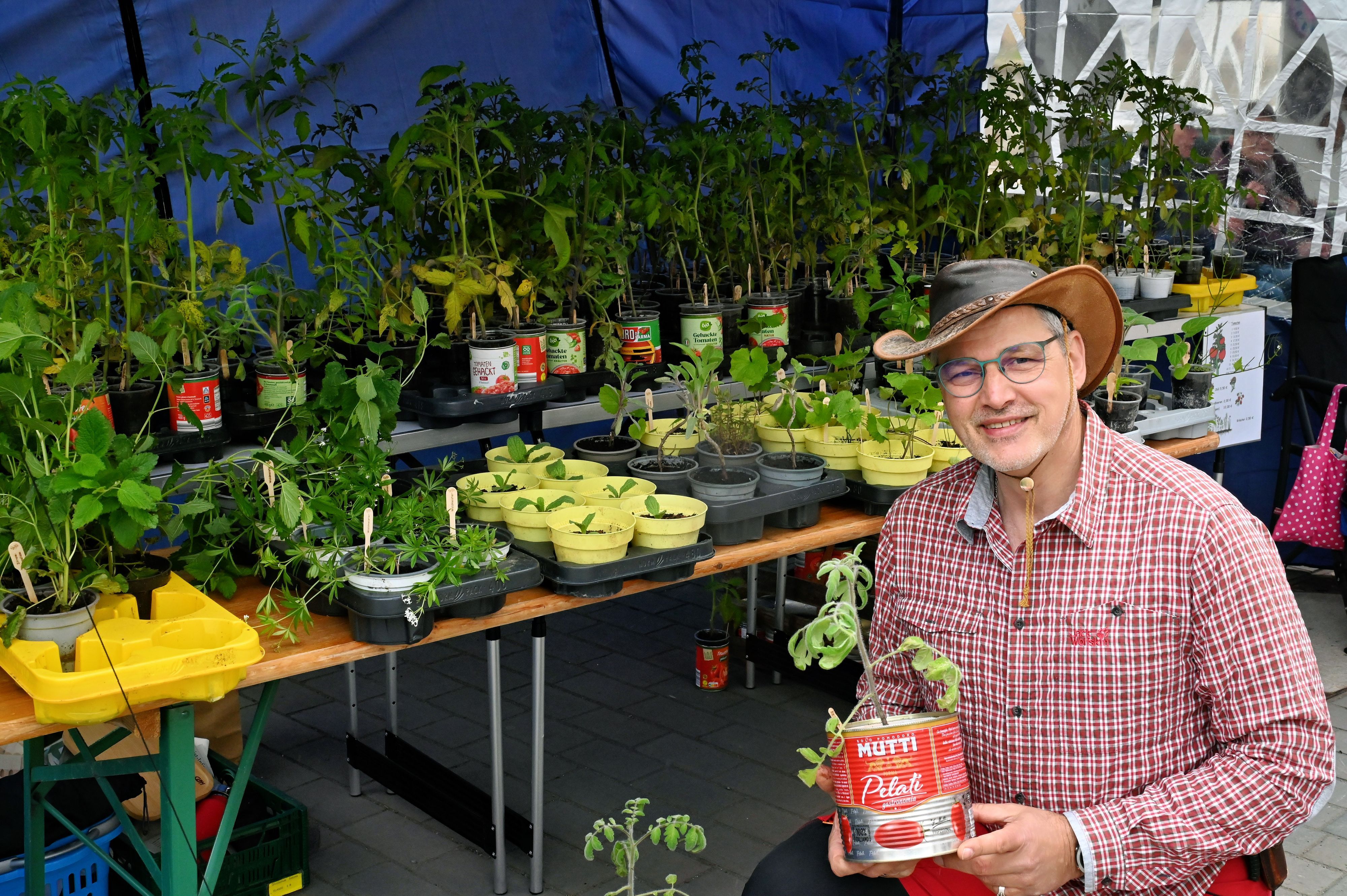 Mike Krüger trägt ein rot-weiß-kariertes Hemd und einen Schlapphut und hält eine Tomatendose in der Hand, aus der eine Tomatenpflanze wächst. Im Hintergrund stehen weitere Pflanzen auf einem Tisch.