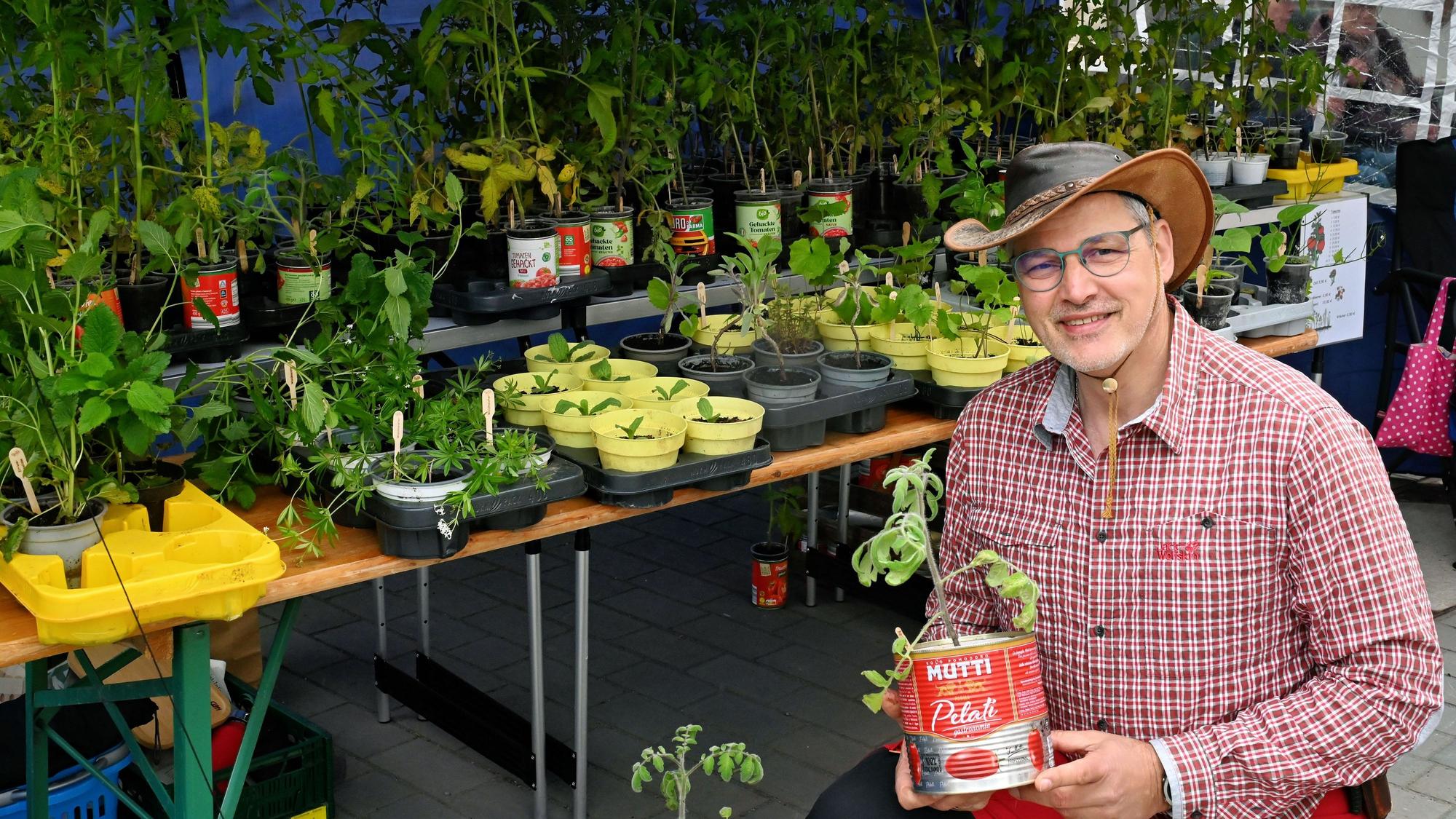 Mike Krüger trägt ein rot-weiß-kariertes Hemd und einen Schlapphut und hält eine Tomatendose in der Hand, aus der eine Tomatenpflanze wächst. Im Hintergrund stehen weitere Pflanzen auf einem Tisch.