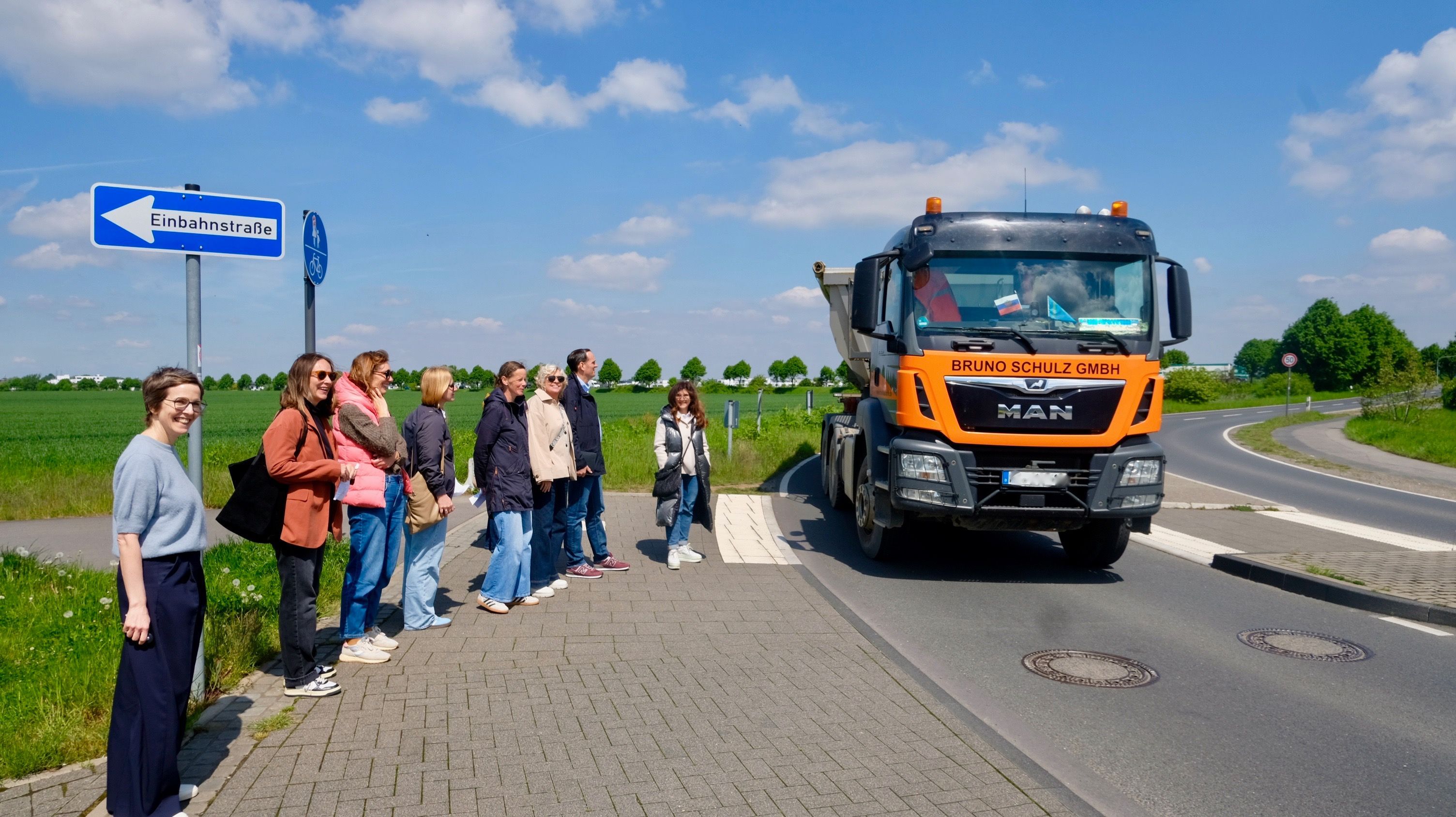 Schulleitung, Eltern und Politiker sowie Politikerinnen warten auf der Aufstellfläche am Fahrbahnrand an der Kurve in der Zusestraße, während ein Lkw durch die Kurve am Gymnasium Zusestraße dicht an der Mittelinsel vorbeifährt.