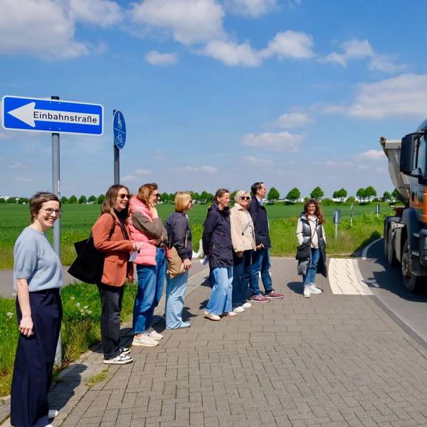 Schulleitung, Eltern und Politiker sowie Politikerinnen warten auf der Aufstellfläche am Fahrbahnrand an der Kurve in der Zusestraße, während ein Lkw durch die Kurve am Gymnasium Zusestraße dicht an der Mittelinsel vorbeifährt.
