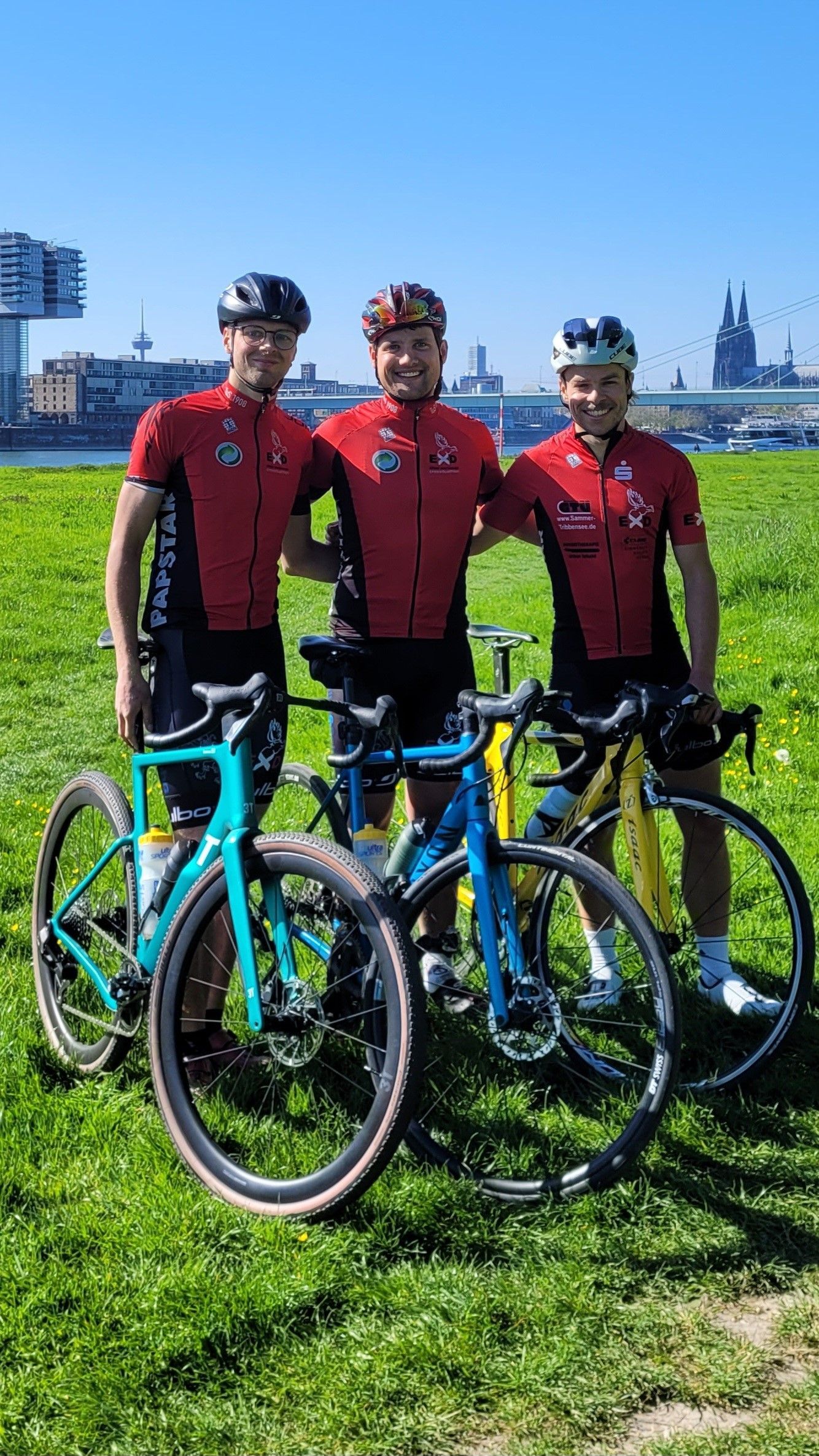 Drei Männer in roten Radfahrtrikots und mit Helmen stehen auf einer Wiese am Rhein vor der Kulisse von Köln.
