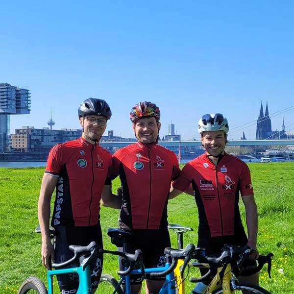 Drei Männer in roten Radfahrtrikots und mit Helmen stehen auf einer Wiese am Rhein vor der Kulisse von Köln.
