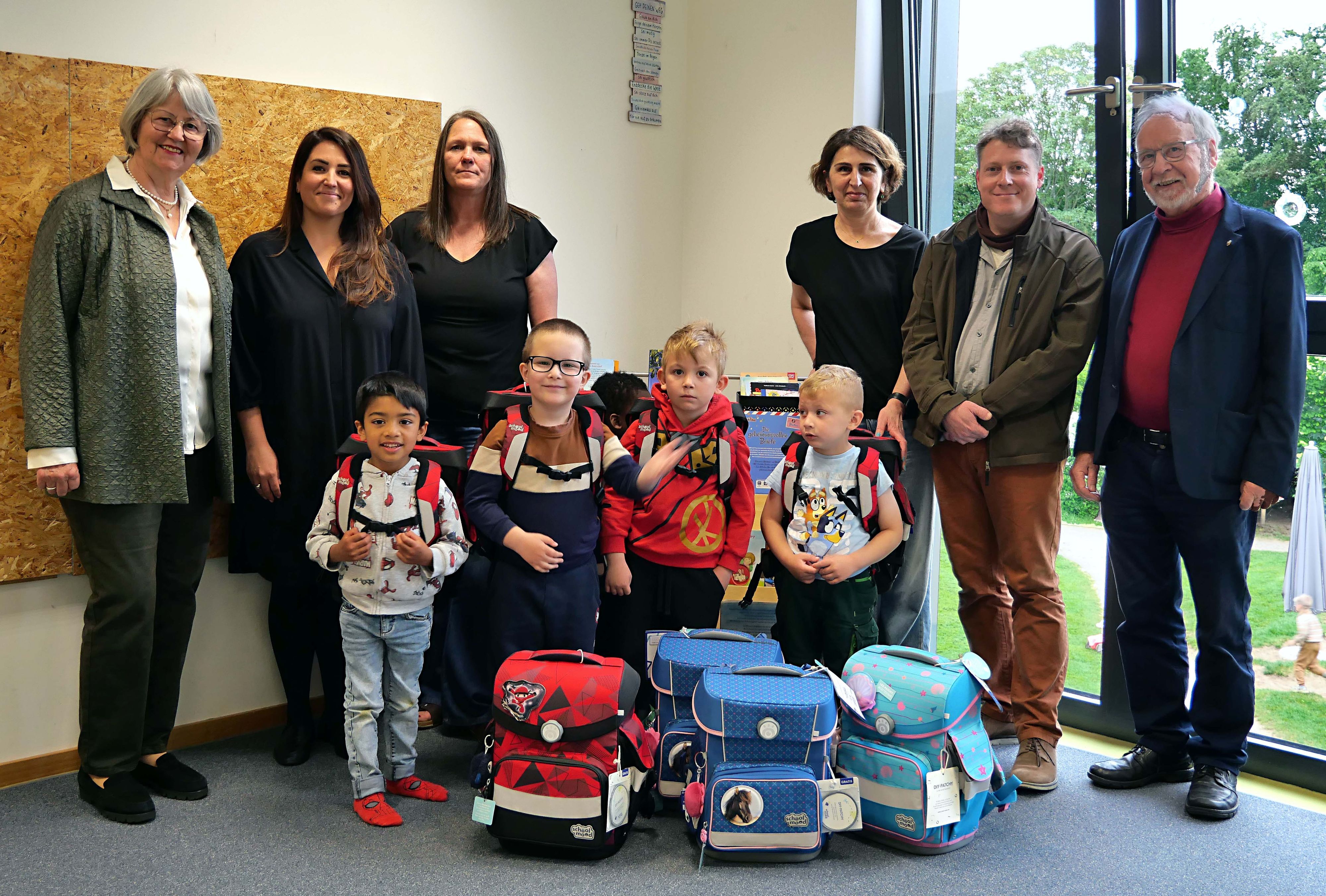 Erwachsene und Kinder mit Schulranzen stehen vor weiteren Ranzen.