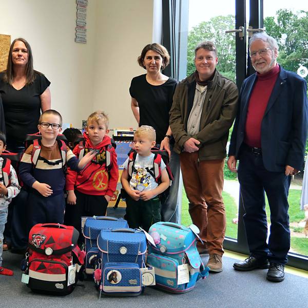 Erwachsene und Kinder mit Schulranzen stehen vor weiteren Ranzen.