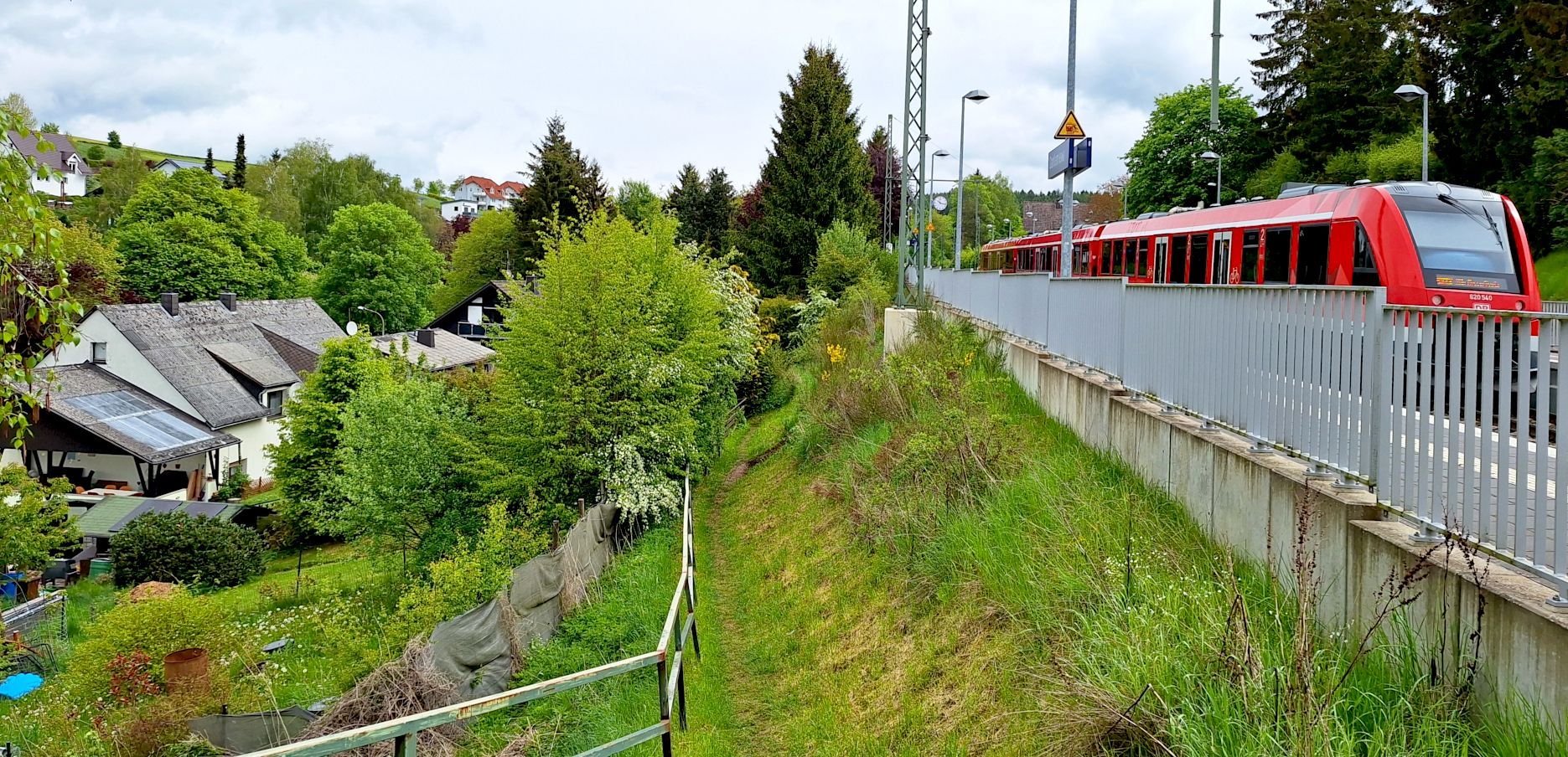 Zwischen den Bahngleisen rechts im Bild, auf denen ein roter Zug fährt, und einem Wohngebiet links führt ein schmaler Pfad über eine Wiese.