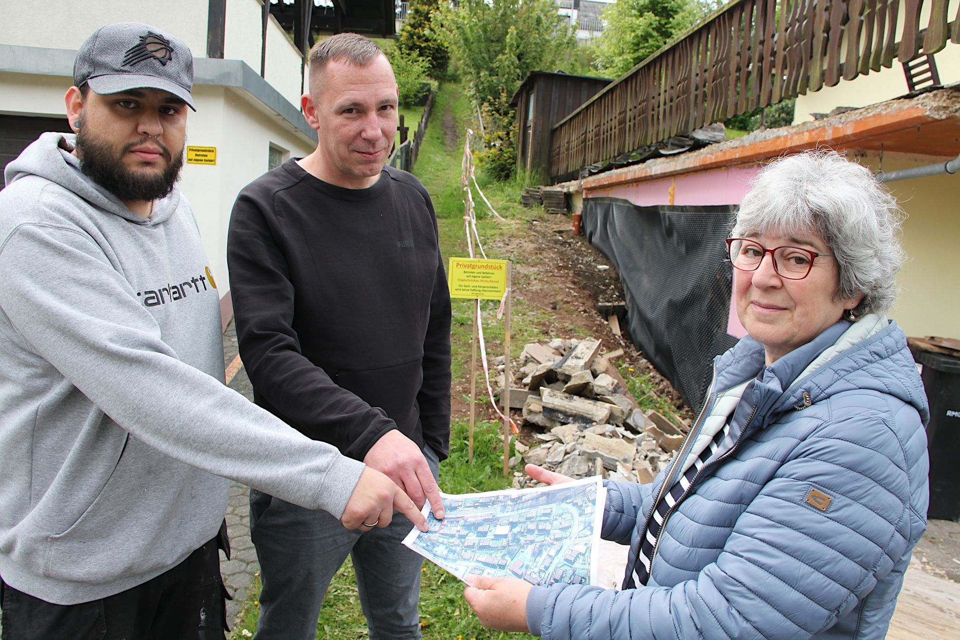 Zwei Männer und eine Frau stehen vor einem schmalen Wiesenweg zusammen. Sie schauen auf ein Luftbild, das die Frau in den Händen hält.