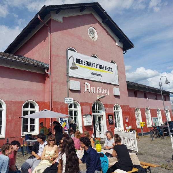 Ein alter Bahnhof, davor sitzen Menschen an Tischen