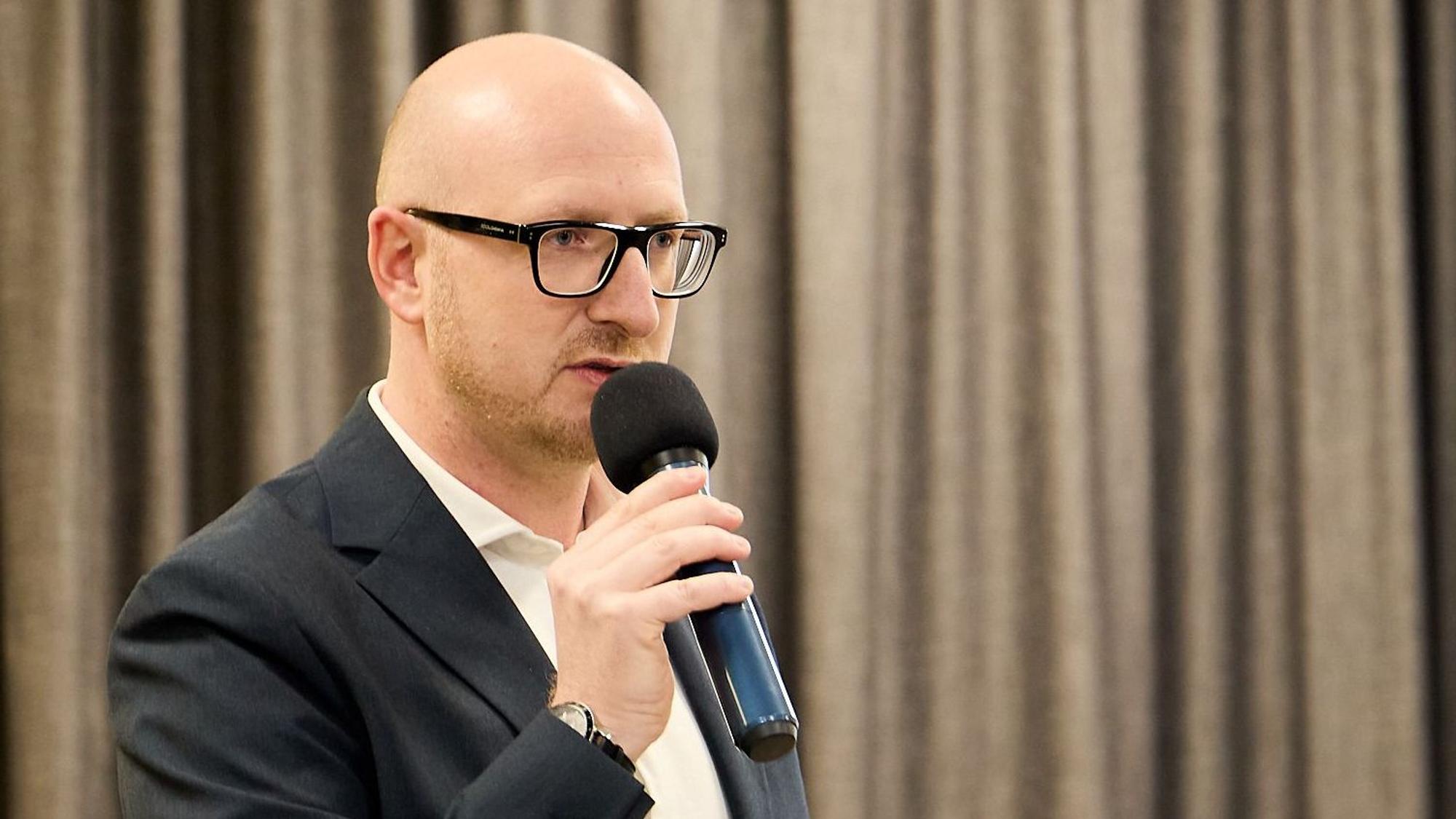 Ingo Pfennings steht mit einem Mikrofon in der Hand vor einem braunen Vorhang.
