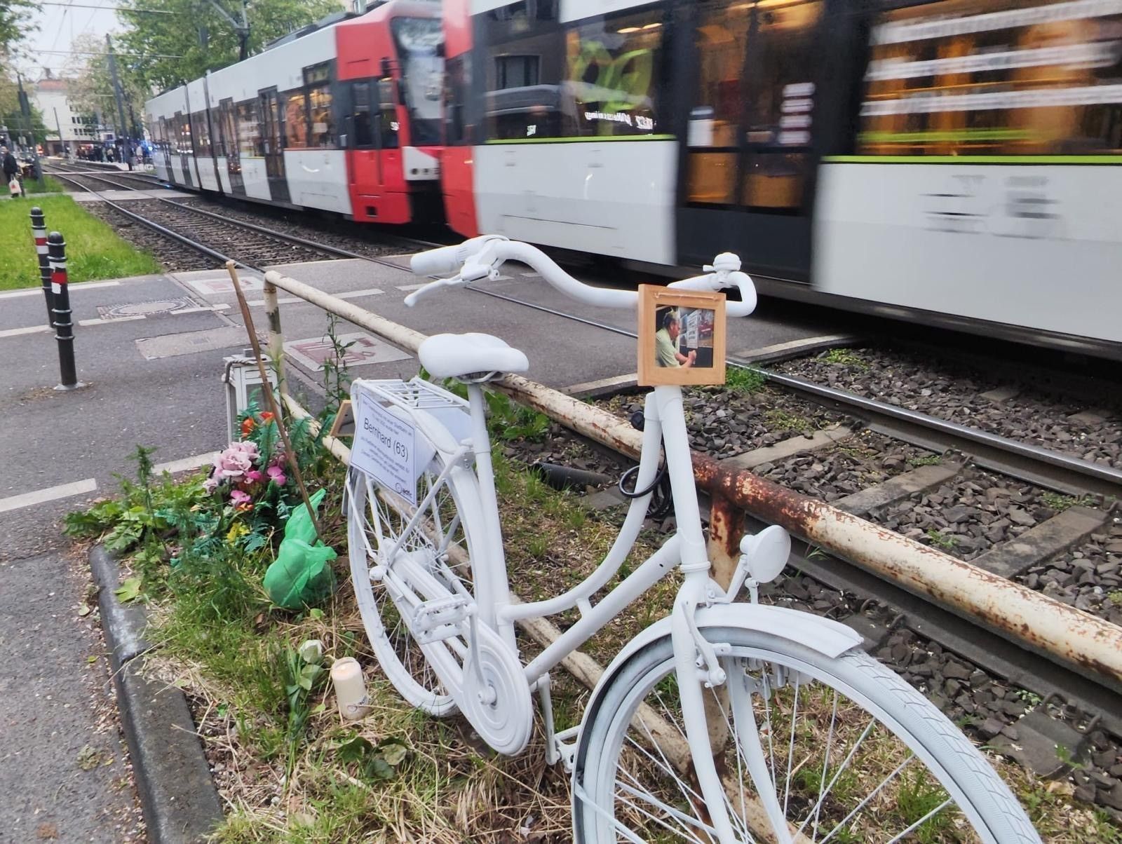Ein weiß lackiertes Fahrrad steht an einem Bahnübergang während ein Zug vorbeirauscht.