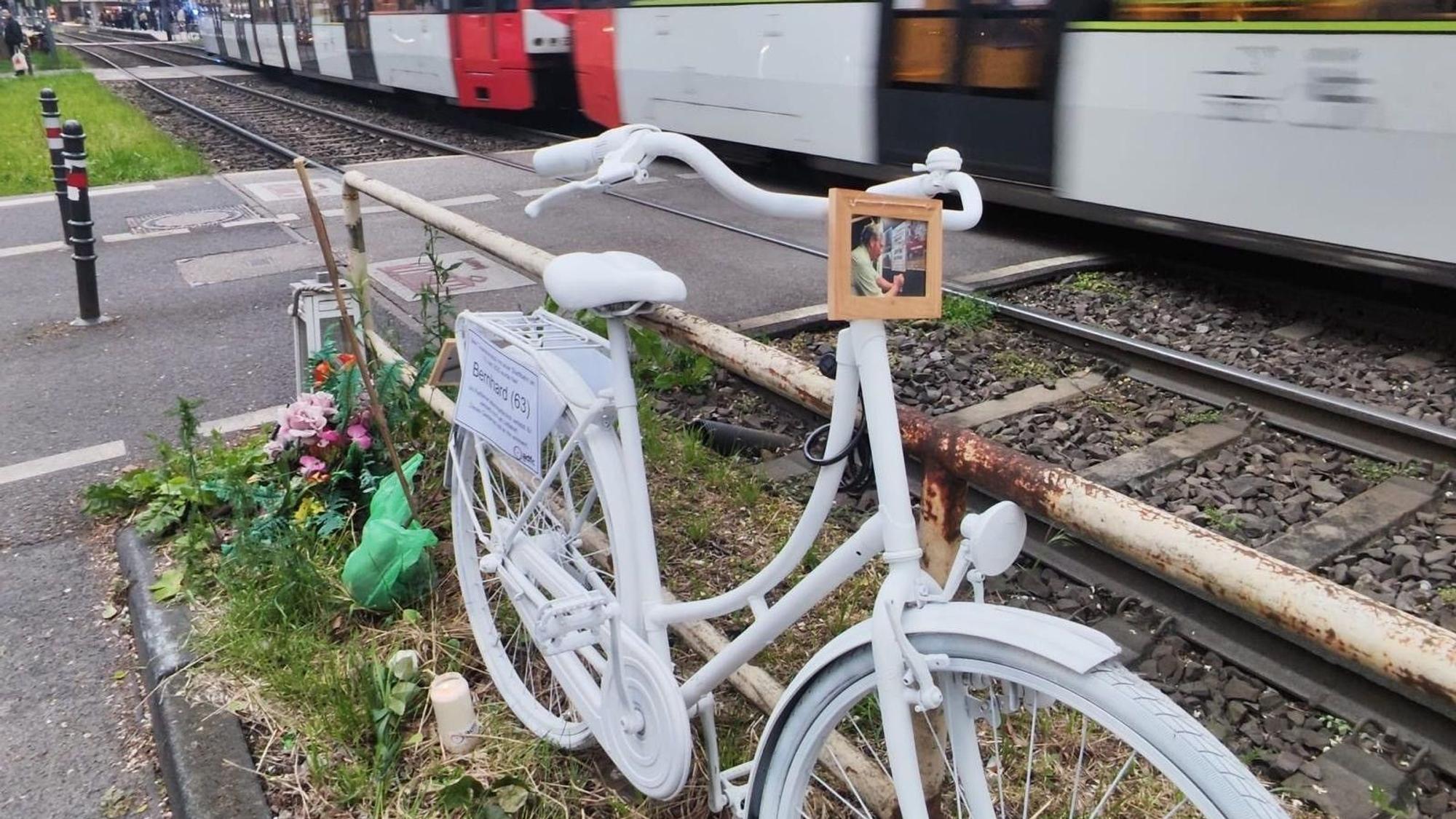 Ein weiß lackiertes Fahrrad steht an einem Bahnübergang während ein Zug vorbeirauscht.