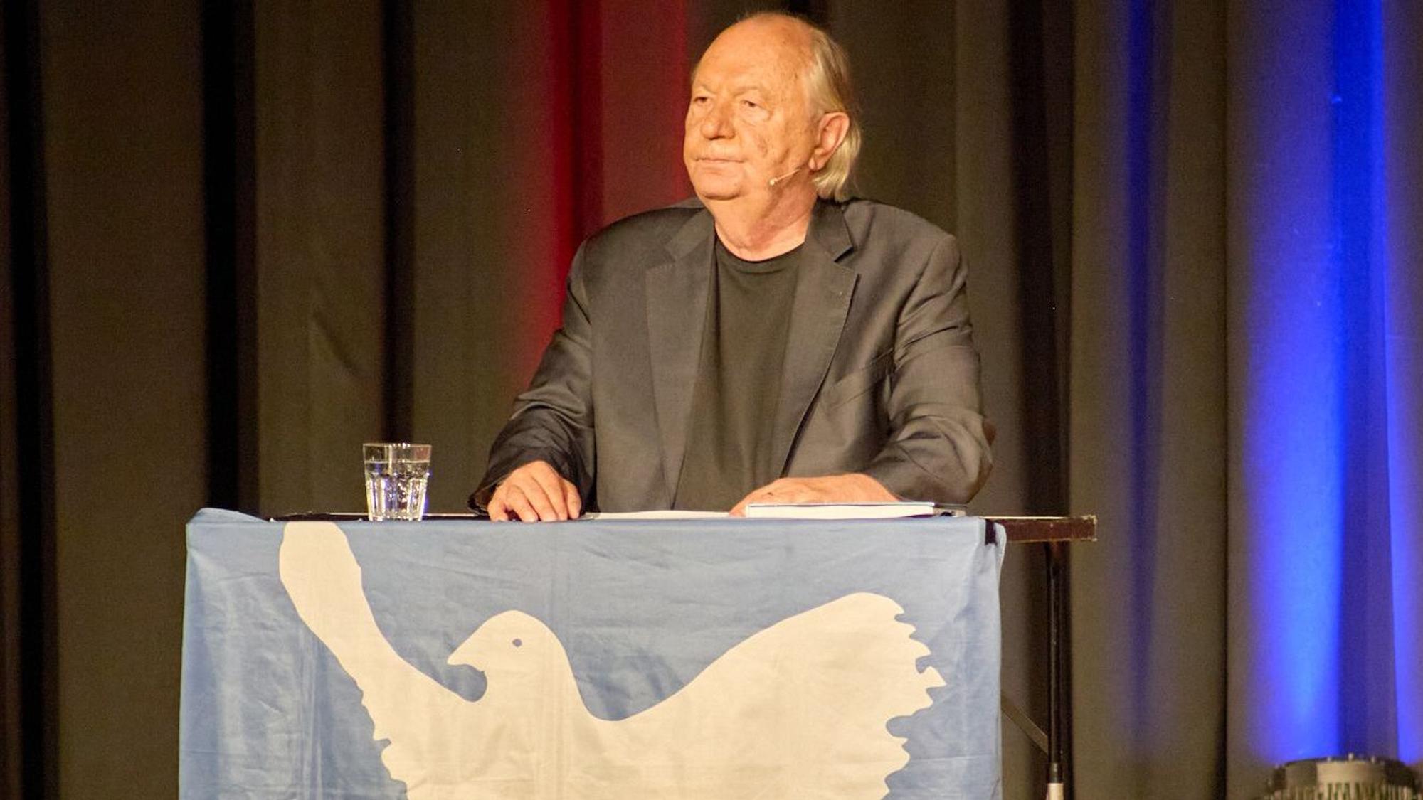 Wilfried Schmickler steht hinter einem Pult, das mit einer Friedenstaube geschmückt ist.