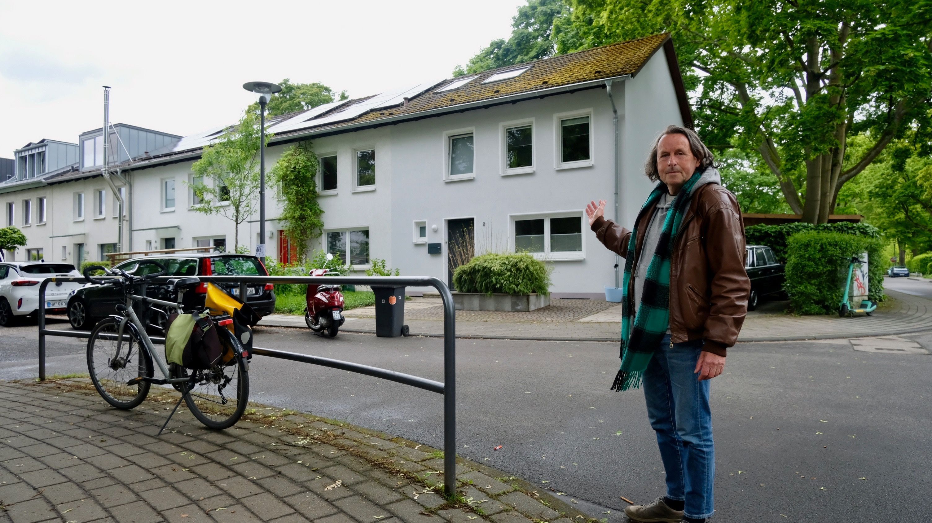 Ein Mann in Lederjacke und Schal steht auf einer Straße. Er weist auf die Reihenhäuser auf der anderen Straßenseite.