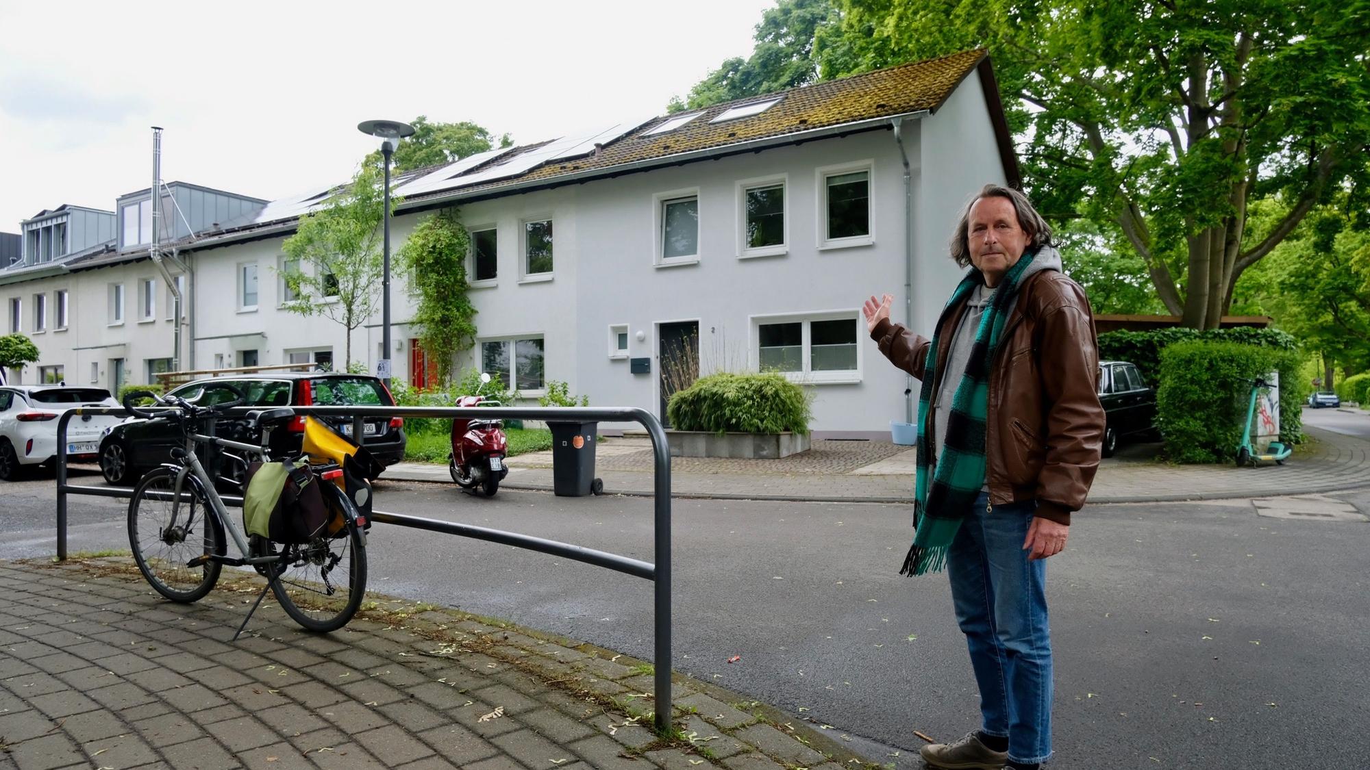 Ein Mann in Lederjacke und Schal steht auf einer Straße. Er weist auf die Reihenhäuser auf der anderen Straßenseite.