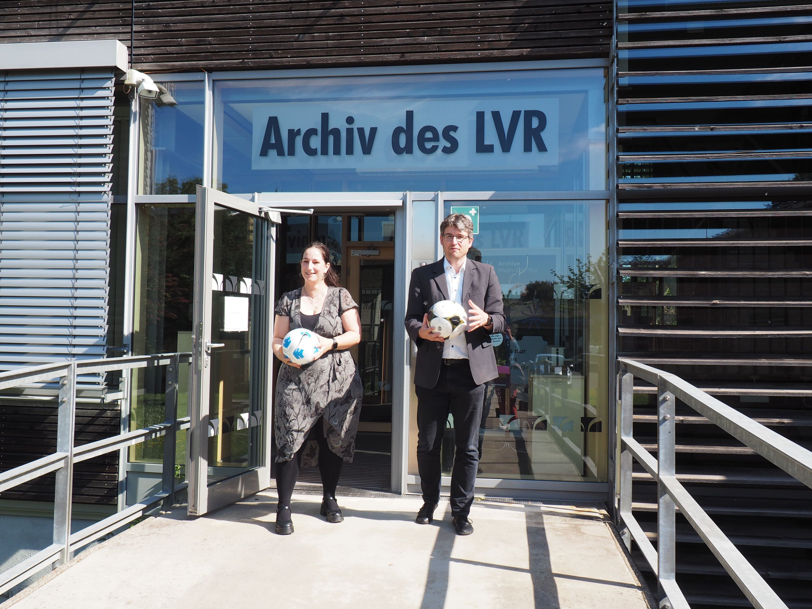 das Foto zeigt eine Frau und einen Mann mit Bällen in den Händen, die aus der Tür des LVR-Archivs kommen.