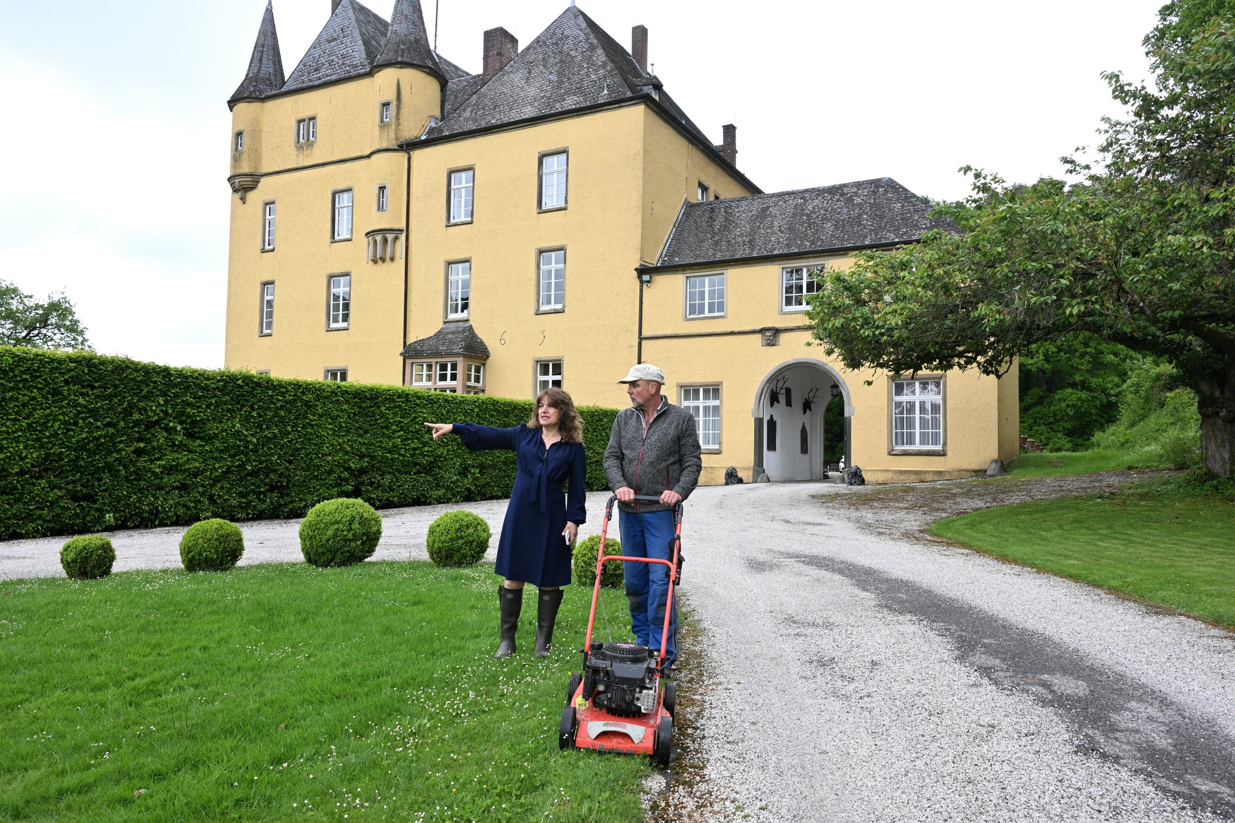 Eine Frau in Kleid und Gummistiefeln steht vor einem Schloss mit gelber Fassade und spricht mit einem Gärtner mit Rasenmäher.