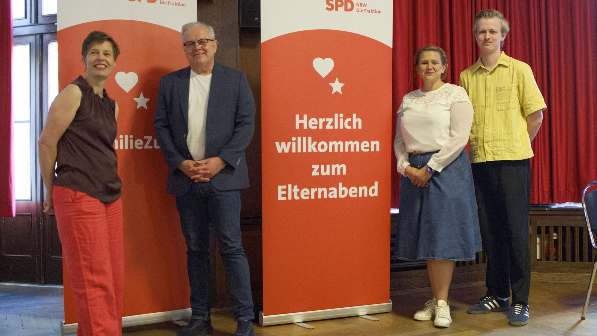 Die Obengenannten stehen neben zwei Plakaten, die auf die Veranstaltung hinweisen.