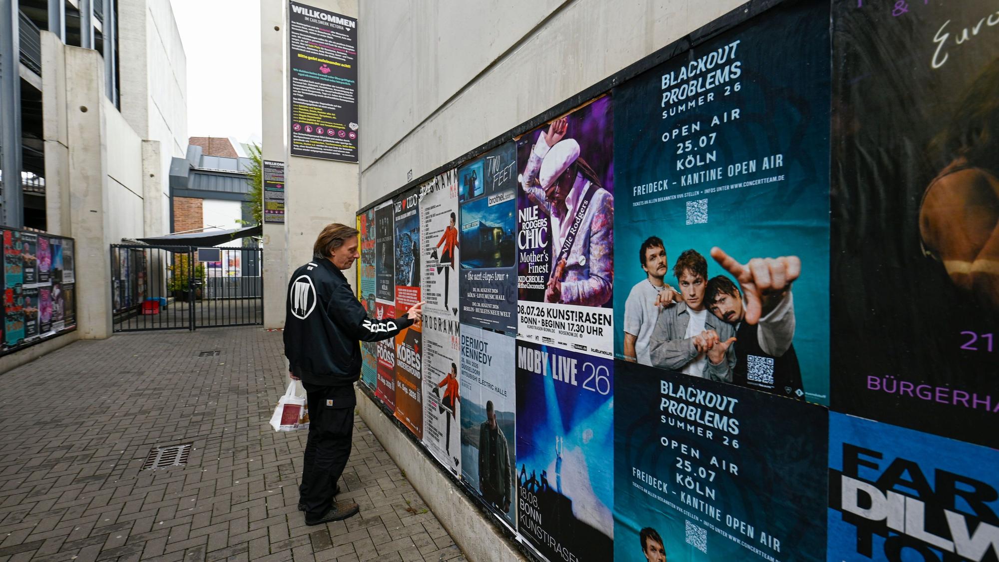 Ein Mann steht vor einer Wand mit Konzert-Plakaten.