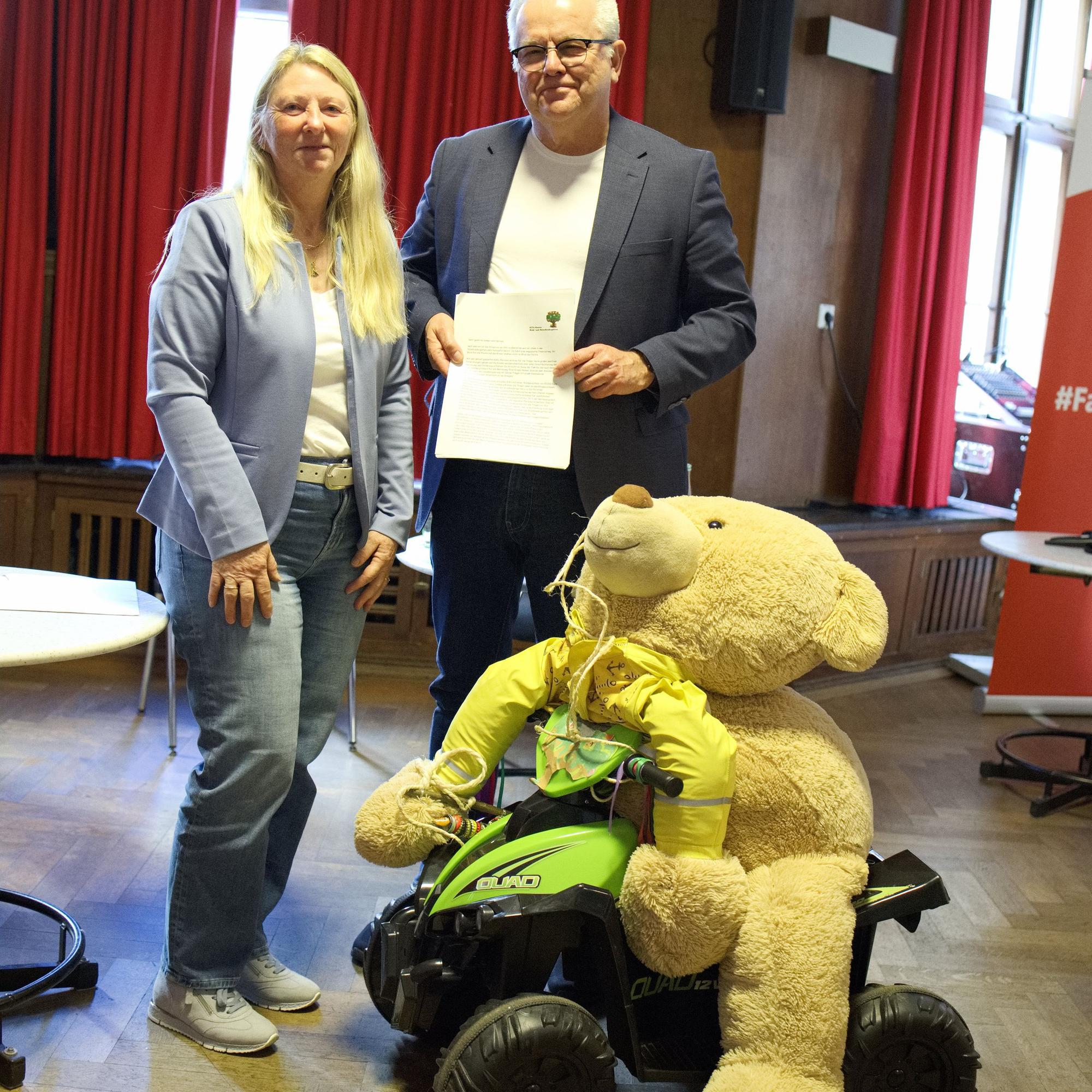 Auf einem großen Spielzeugtraktor sitzt ein großer Teddybär. Dahinter stehen ein Mann und eine Frau.