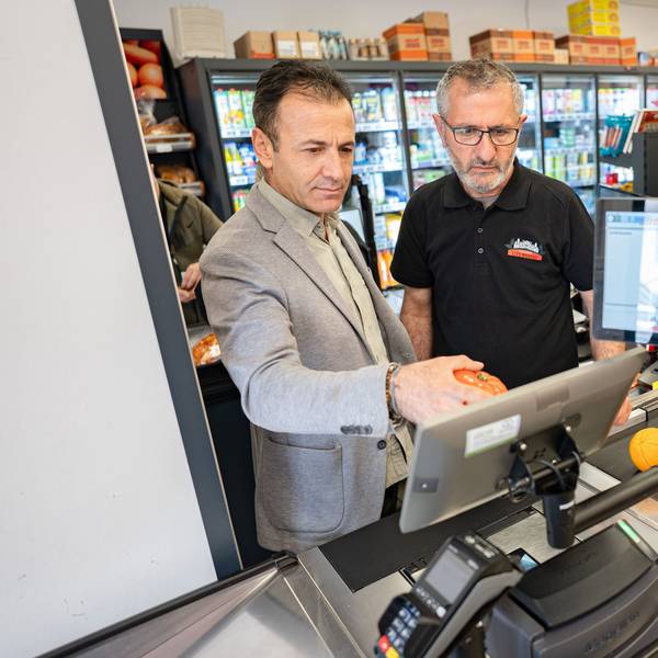 Der Männer stehen auf der Kassierer-Seite an einer Supermarkt-Kasse und bedienen das Display. Auf dem Band liegen Obst und Gemüse