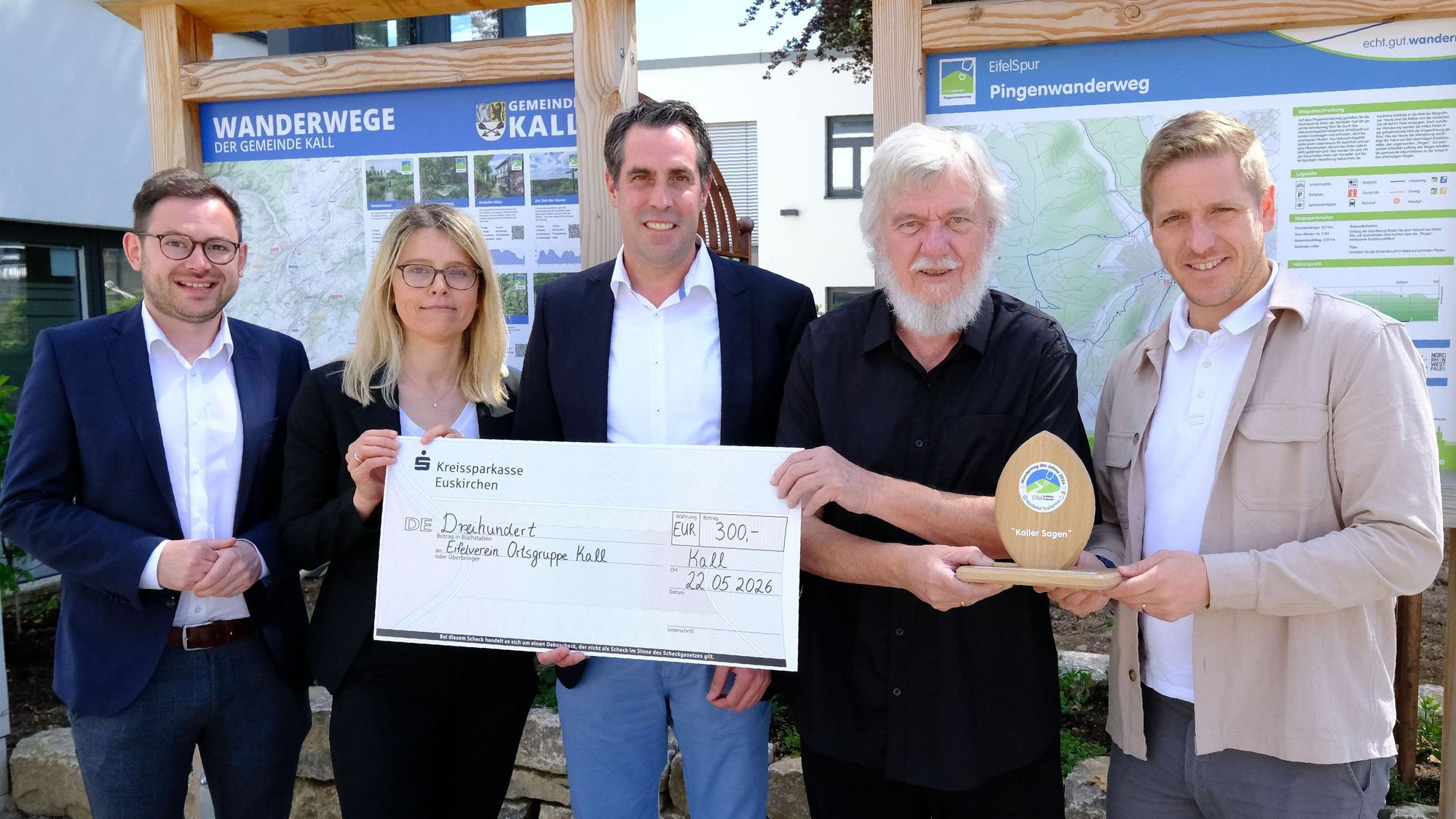 Die Genannten stehen mit einem überdimensionierten Scheck und einer Sieger-Trophäe vor zwei Wander-Infotafeln am Kaller Rathaus.