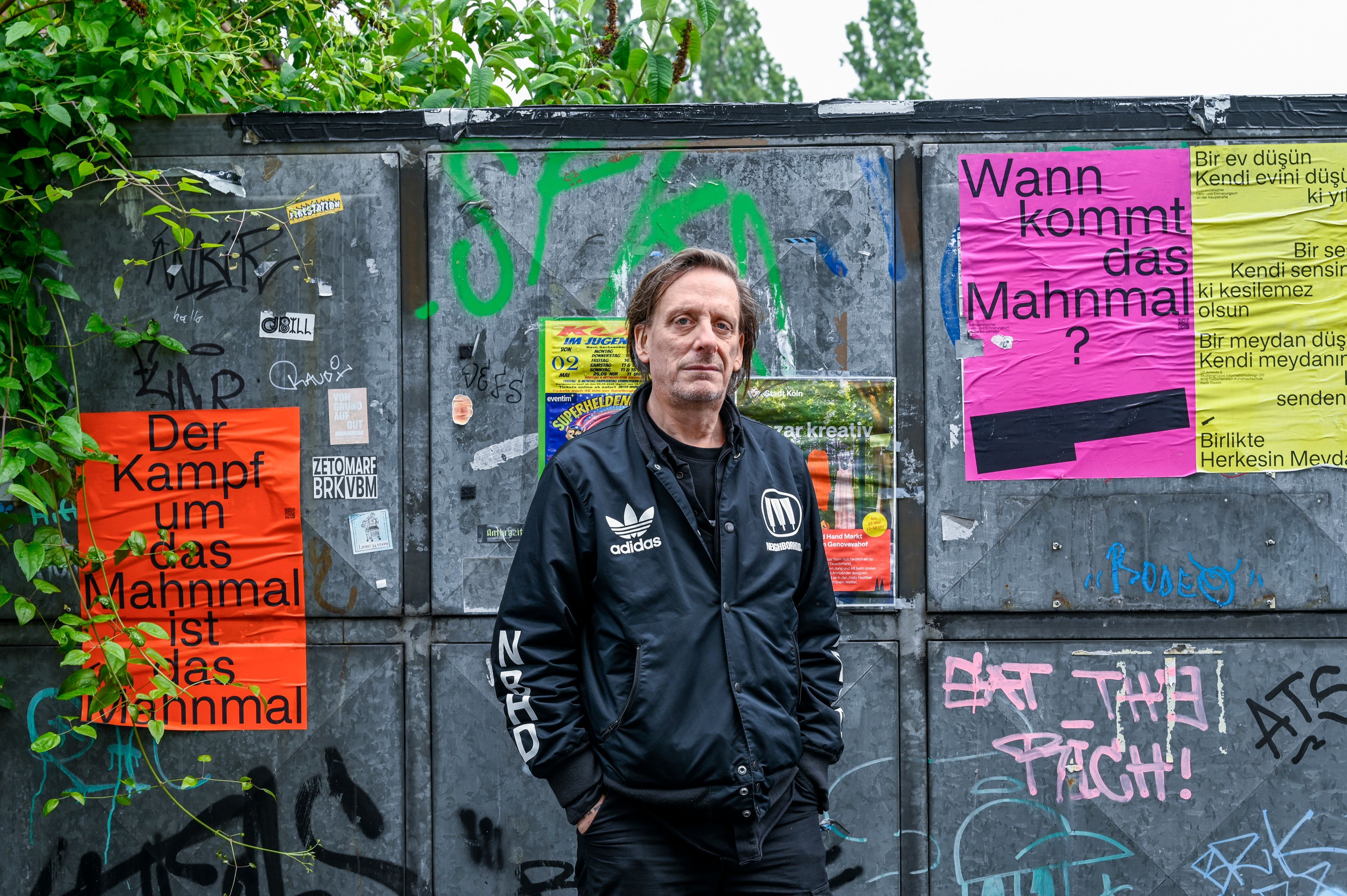 Ein Mann in schwarzer Adidas-Jacke steht vor einem Metalltor, auf dem Plakate kleben, die das fehlende Mahnmal zum Thema haben.